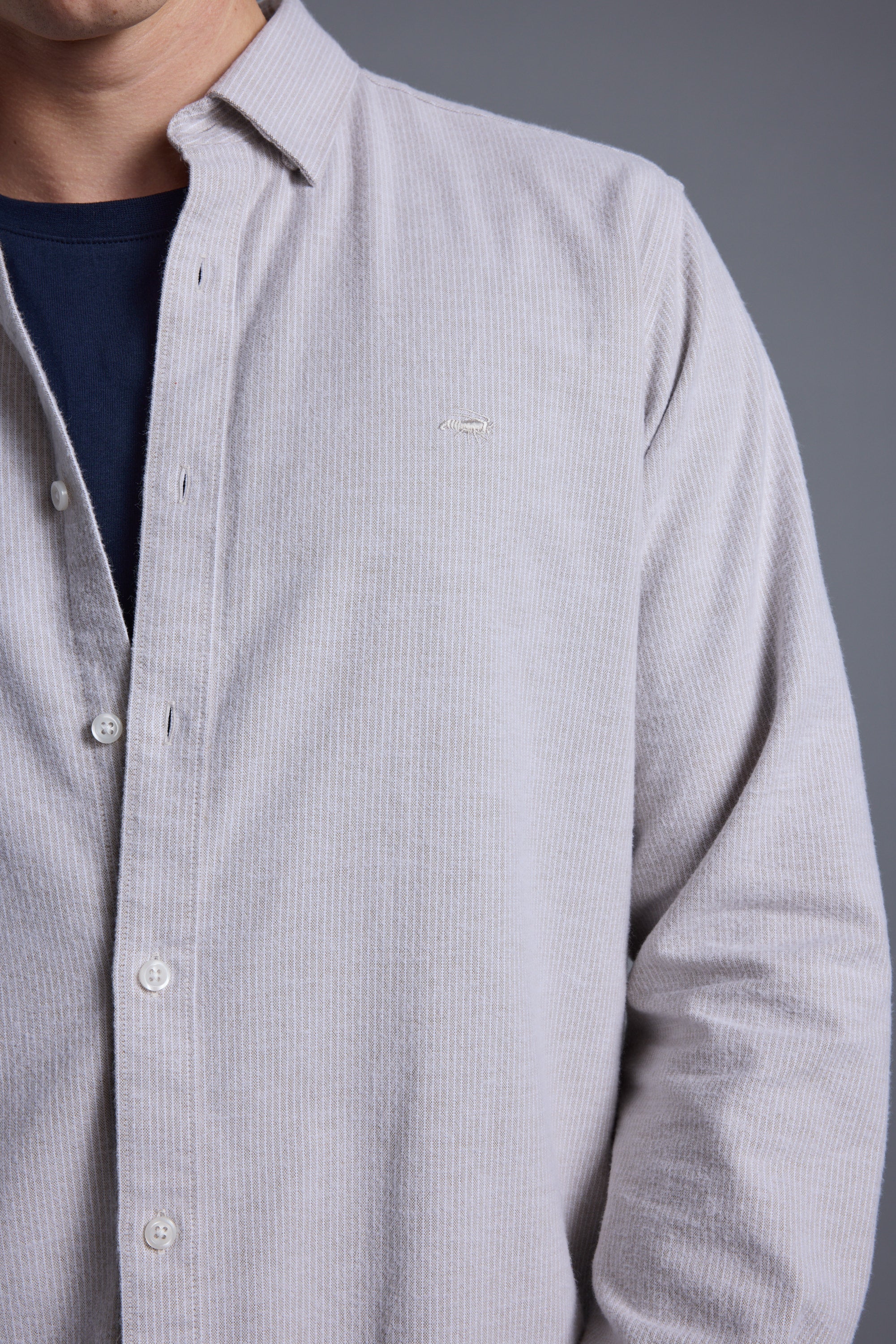 Wearing the Barkers Maynard Oxford Stripe Shirt—light gray with vertical stripes, made from responsibly sourced cotton—over a dark blue T-shirt. Shown from shoulders to waist against a plain gray background; face partially out of frame.