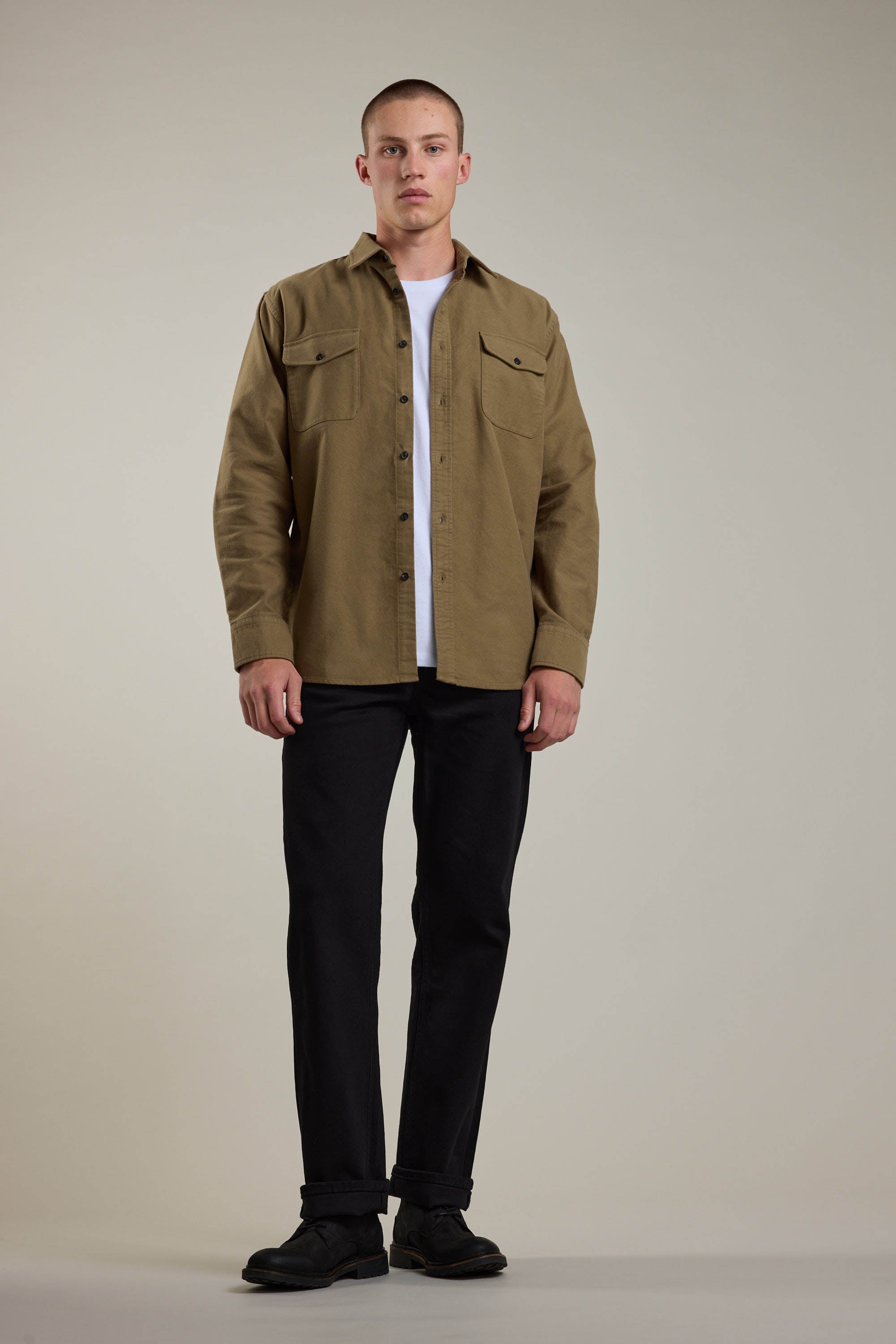 A man stands facing forward in the Barkers Tayler Moleskin Overshirt—a tan, suede-textured cotton layer—worn over a white t-shirt with black pants and shoes, set against a plain light background.