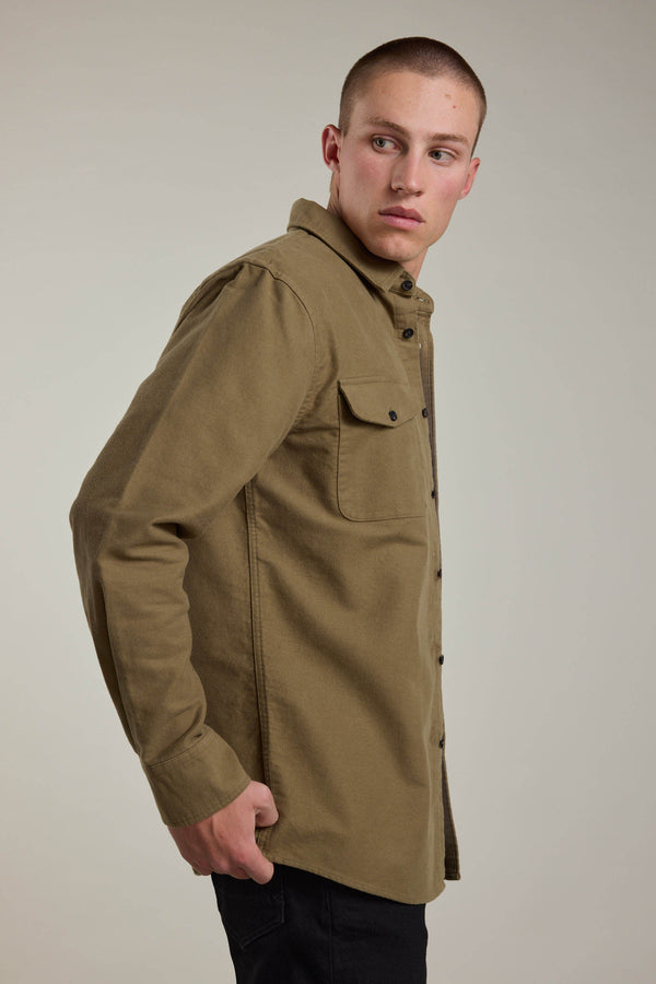 A young man with short hair wears the Barkers Tayler Moleskin Overshirt in olive green and black pants, standing against a plain light background while looking over his left shoulder with a neutral expression.