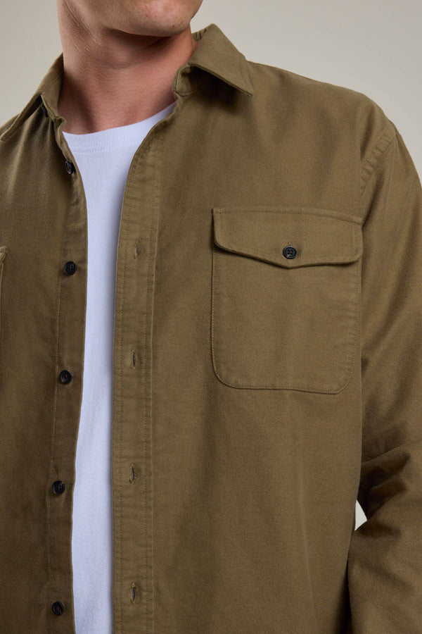 A person wears the Barkers Tayler Moleskin Overshirt in olive green, crafted from responsibly sourced cotton with a buttoned chest pocket and black buttons, over a white t-shirt. The person's head is partially out of frame.