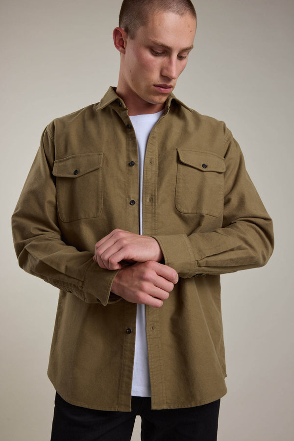 A person wearing the Barkers Tayler Moleskin Overshirt in olive-green over a white T-shirt stands against a plain background, looking down while fastening their sleeve cuff.