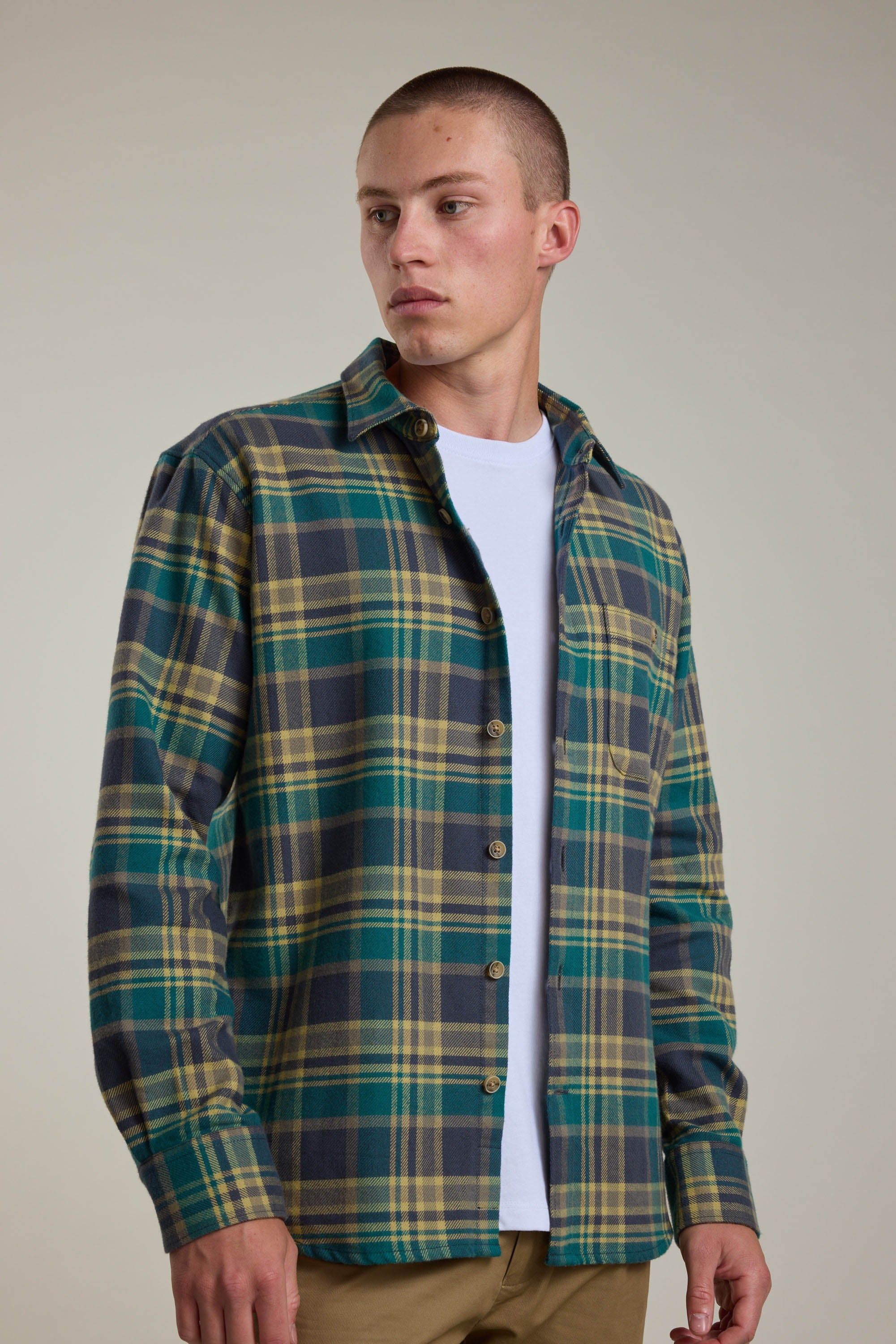 A young man with short hair wears the Barkers Wallis Check Overshirt in green, yellow, and navy plaid over a white tee and tan pants, standing against a plain light background. He looks left—this shirt is a true men's wardrobe essential.
