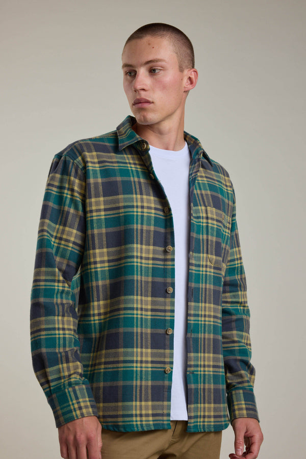 A young man with short hair wears the Barkers Wallis Check Overshirt in green, yellow, and navy plaid over a white tee and tan pants, standing against a plain light background. He looks left—this shirt is a true men's wardrobe essential.