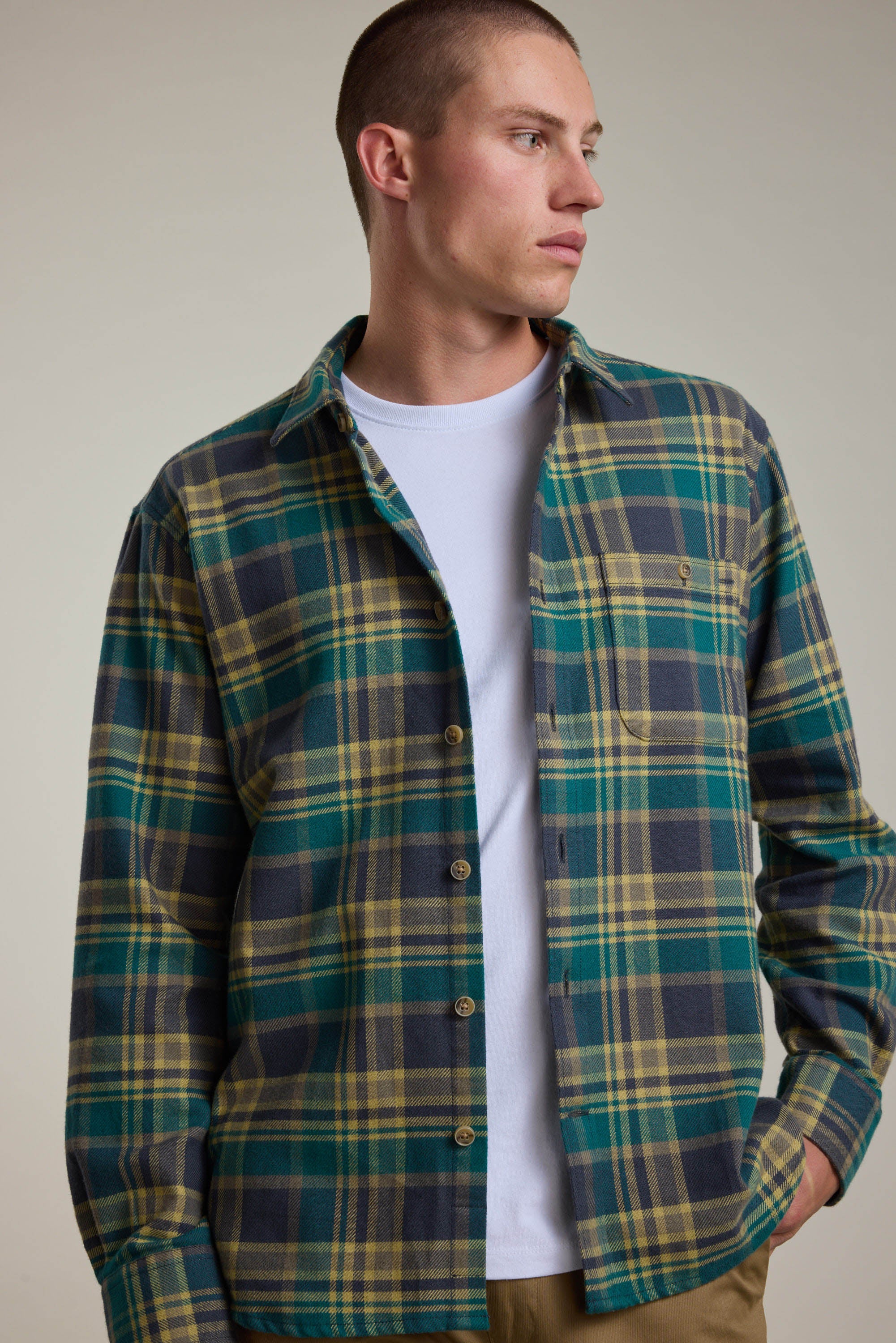 A man in the Barkers Wallis Check Overshirt—green, yellow, and gray checks—layered over a white tee stands with one hand in his pocket, glancing sideways against a plain background.