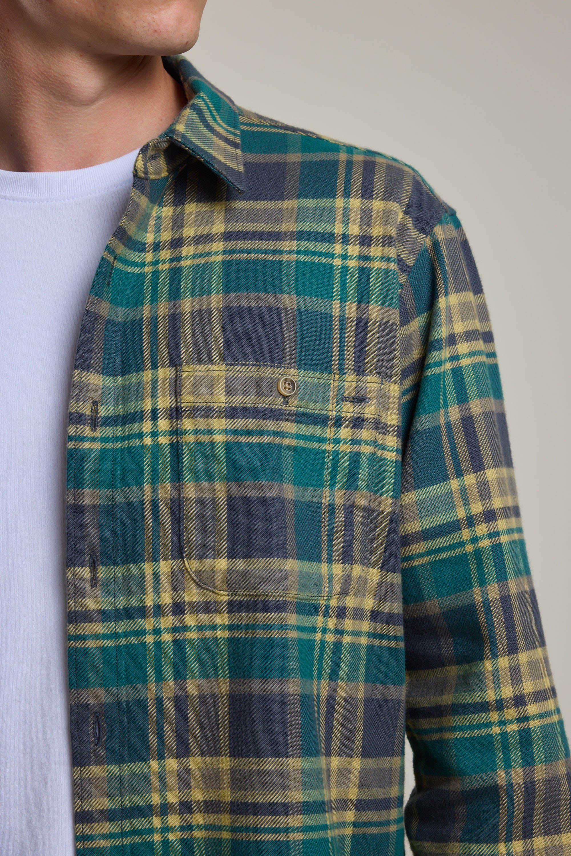 A man wears the Barkers Wallis Check Overshirt—a green, yellow, and gray plaid flannel—over a white tee. The photo shows his upper torso and face, highlighting this men's wardrobe essential’s pattern and front pocket.