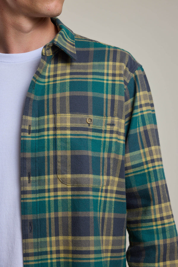 A man wears the Barkers Wallis Check Overshirt—a green, yellow, and gray plaid flannel—over a white tee. The photo shows his upper torso and face, highlighting this men's wardrobe essential’s pattern and front pocket.