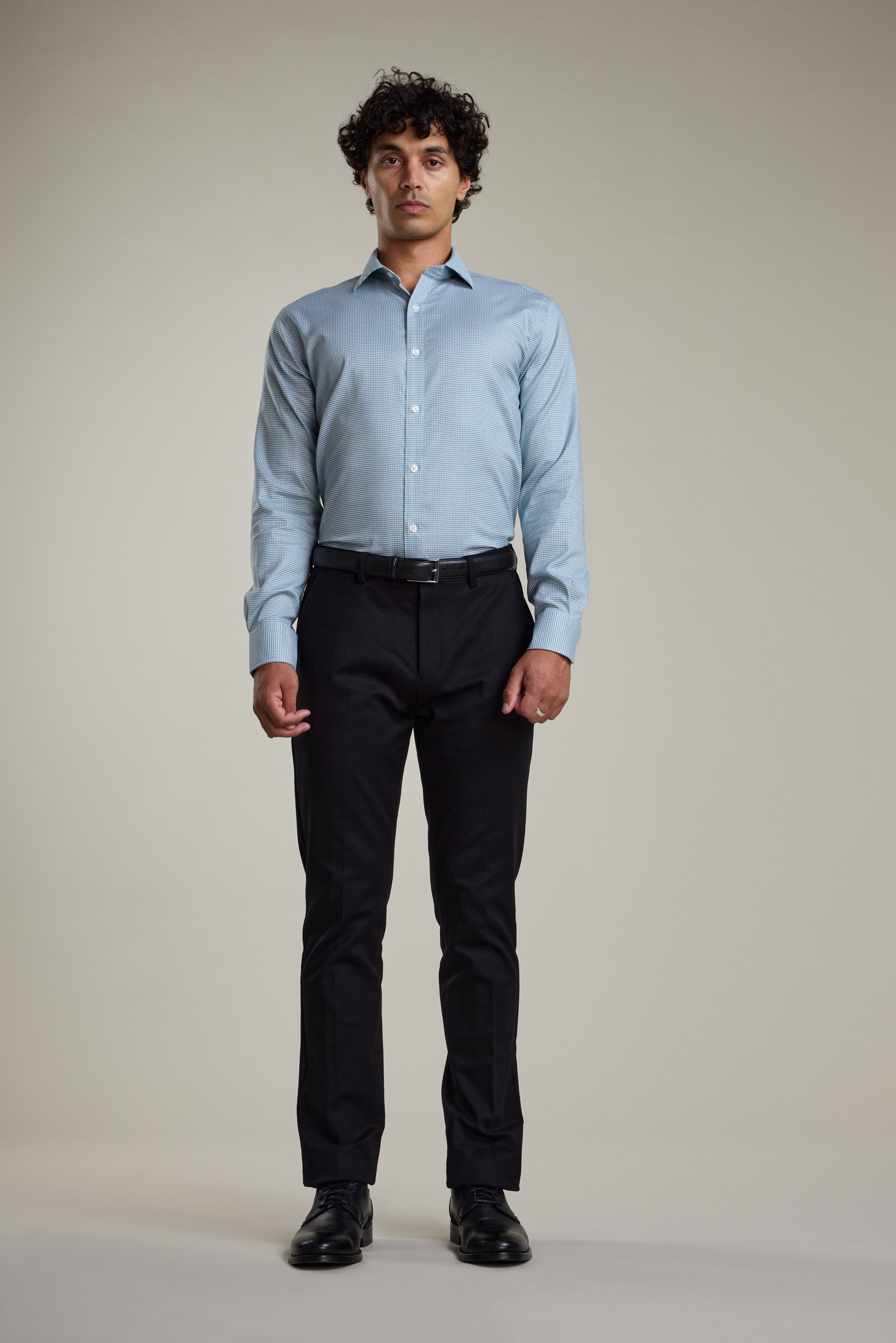 A man with curly hair stands facing forward, wearing a Barkers Zione Business Shirt in light blue, black dress pants, and black shoes against a plain beige background.