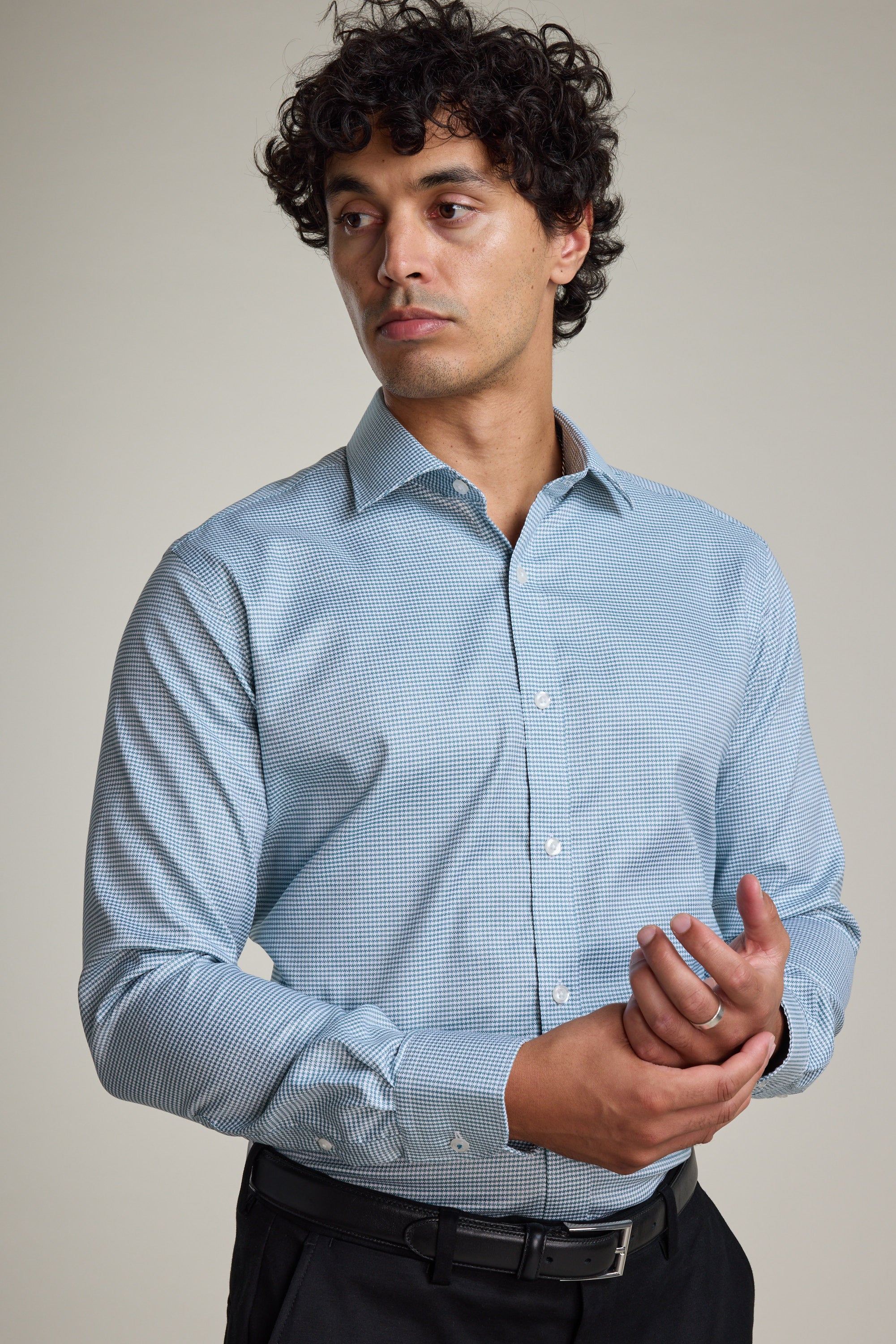A man with curly dark hair wears the Barkers Zione Business Shirt, a light blue houndstooth-patterned shirt, and black pants. He stands against a plain background, looking left while adjusting his right sleeve with his left hand.