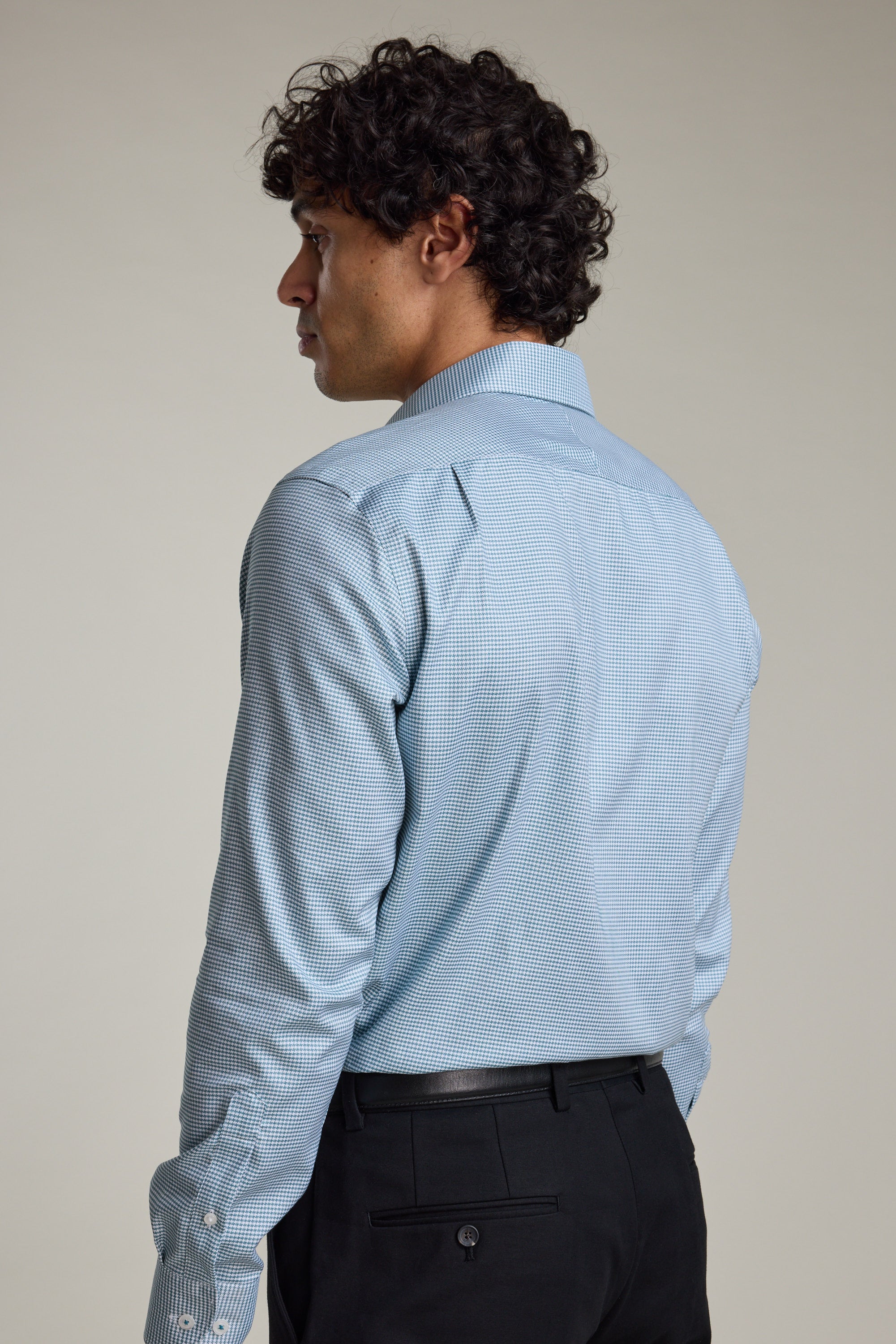 A man with curly dark hair is shown from the back, wearing a light blue Barkers Zione Business Shirt and black trousers, standing against a plain light background.