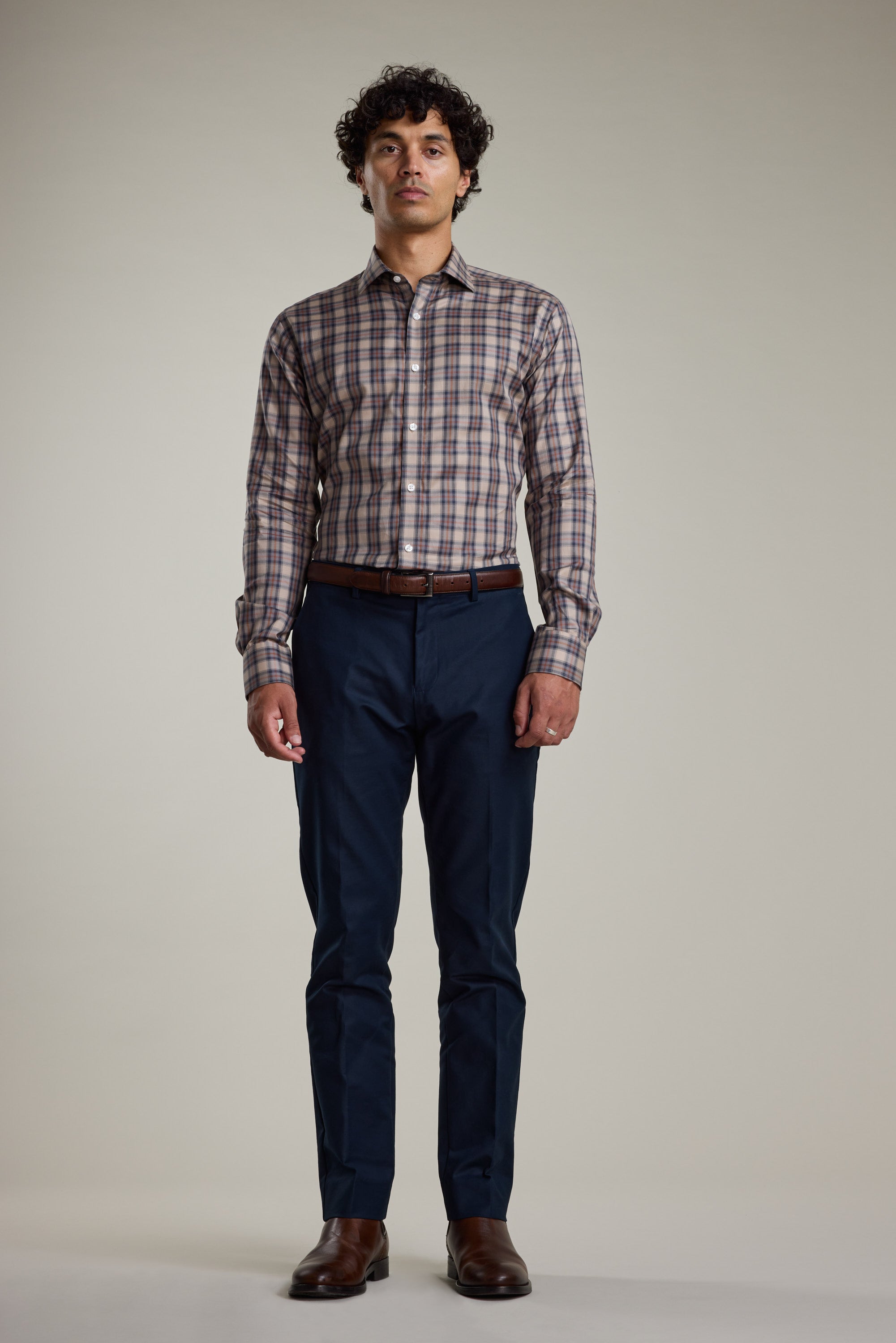 A man faces forward wearing the Barkers Norco Check Business Shirt, a tailored fit brown plaid button-up, paired with dark blue pants, a brown belt, and brown shoes against a plain light background.