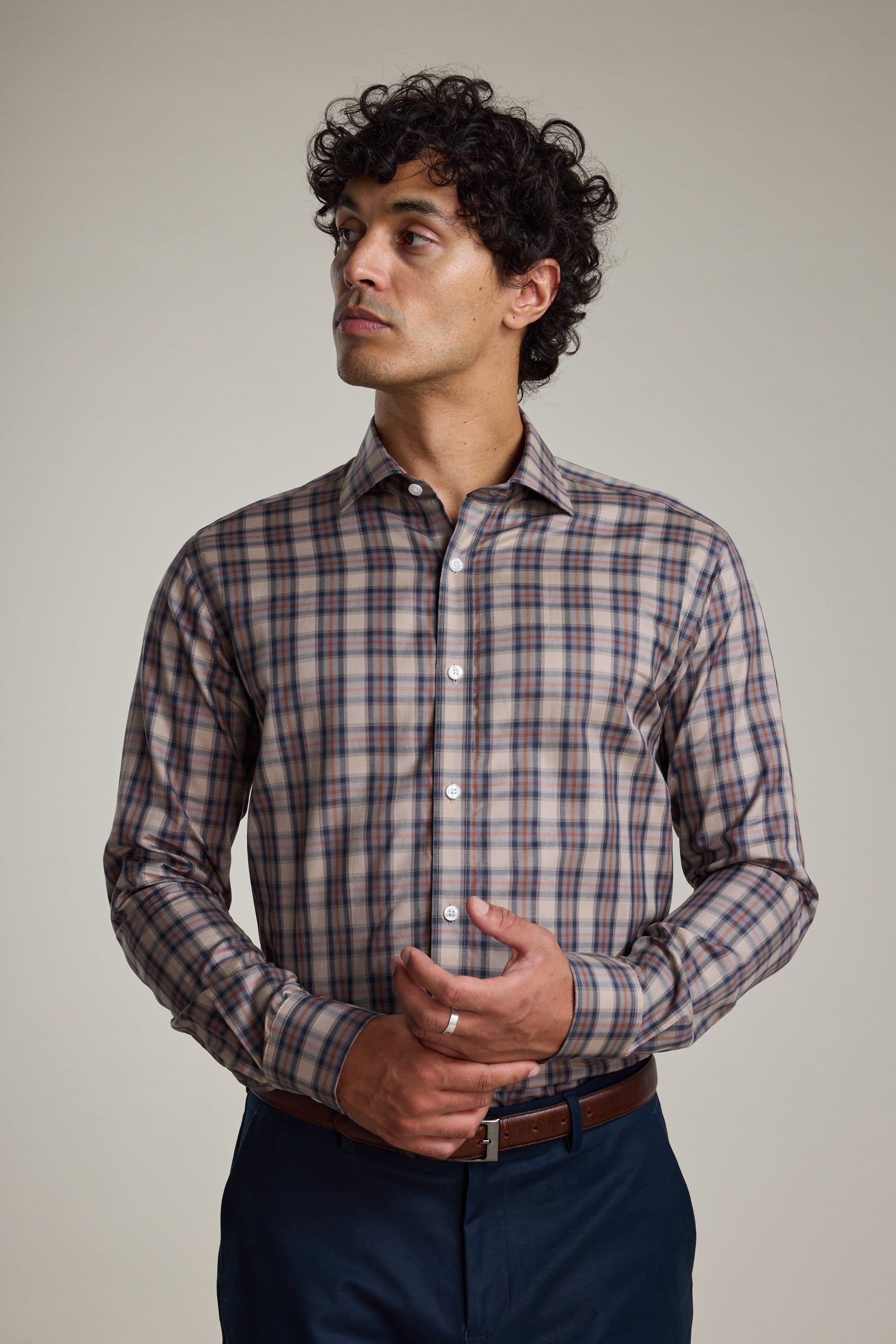 A man with curly dark hair wears the Barkers Norco Check Business Shirt, a wrinkle resistant, tailored fit plaid shirt in beige, brown, and blue tones. He pairs it with dark pants and looks to the side with hands loosely clasped.