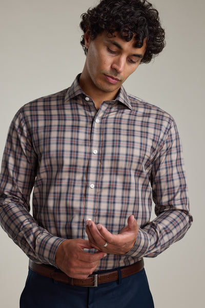 A man with curly hair models the Barkers Norco Check Business Shirt—a brown and blue plaid tailored-fit style—paired with dark pants, as he looks down and touches his left hand with his right against a plain background.