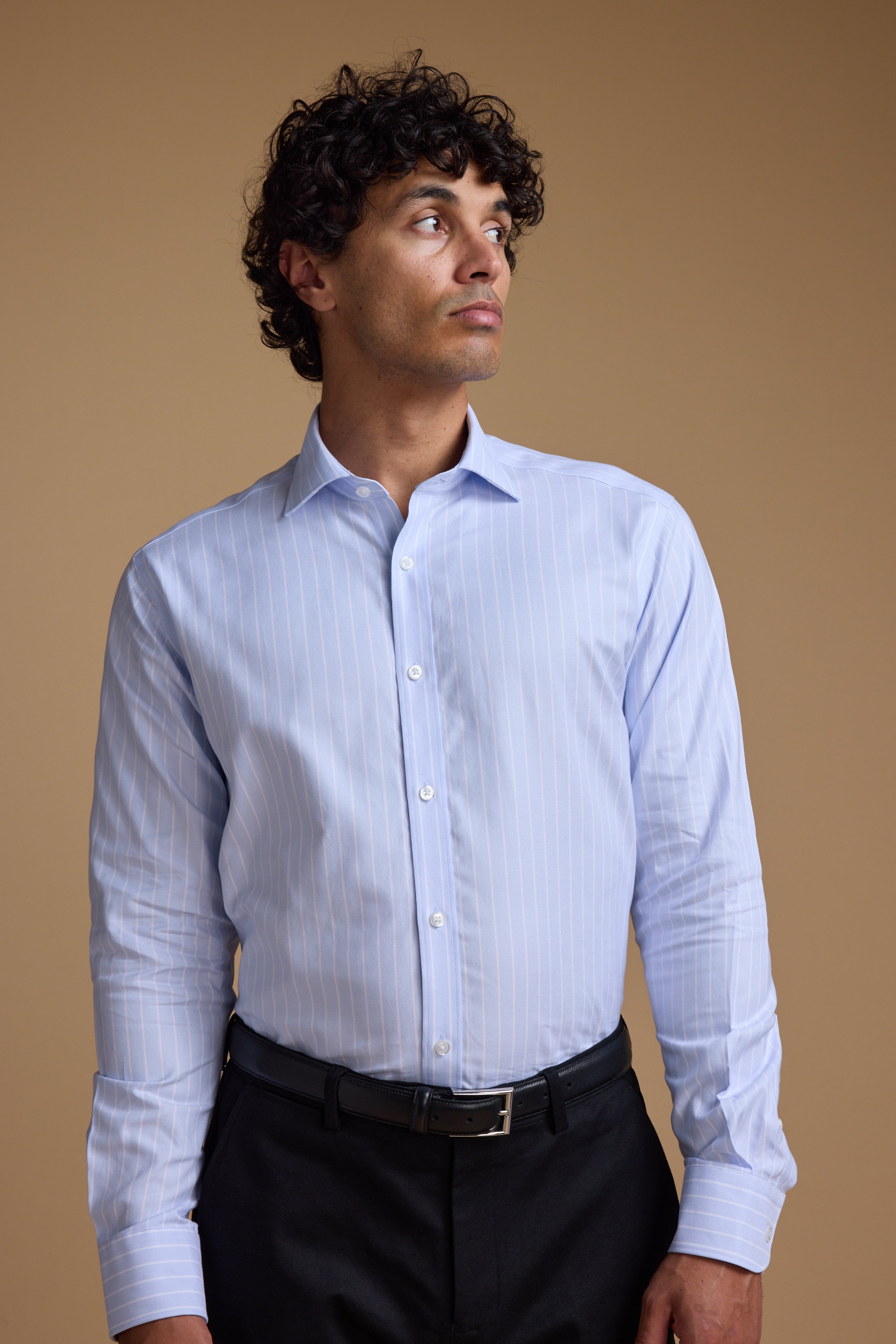 A man with curly hair wears the Barkers Goodall Stripe Business Shirt in light blue and black pants, standing against a plain beige background and looking slightly to his left.