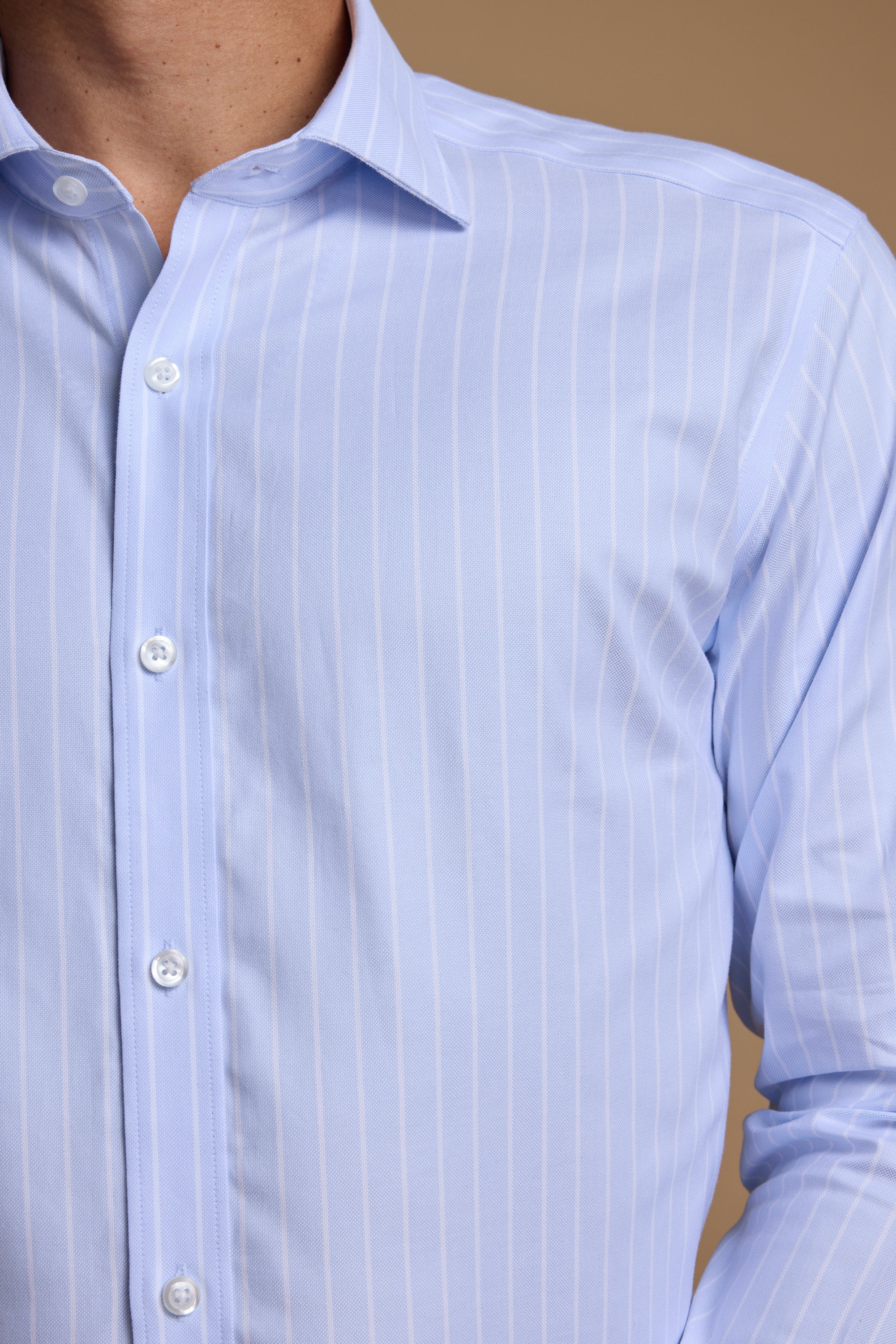 Close-up of a person wearing the Barkers Goodall Stripe Business Shirt, a light blue, long-sleeve wrinkle-free shirt with thin white stripes and white buttons, shown against a beige background with only the torso and neck visible.