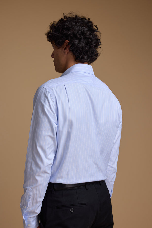 A man with curly dark hair, wearing the Barkers Goodall Stripe Business Shirt in light blue and black pants, stands facing away from the camera against a plain beige background.