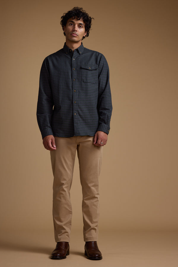 A man with curly hair stands against a beige background, wearing the Barkers Starle Check Shirt made from responsibly sourced cotton, tan pants, and brown boots. His arms rest at his sides as he looks slightly upward.