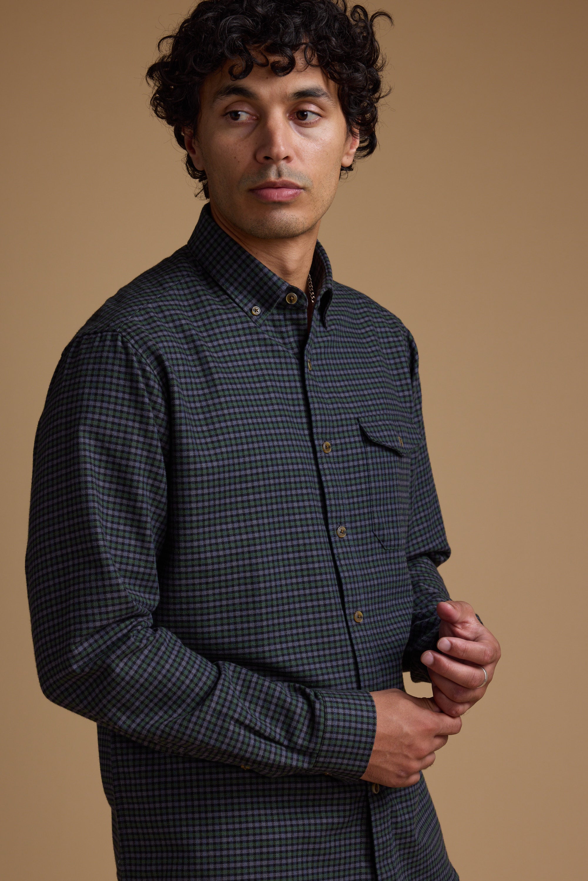 A man with curly brown hair wears the Barkers Starle Check Shirt, a vintage-style design made from responsibly sourced cotton, standing against a plain beige background and looking slightly to his left with a neutral expression.