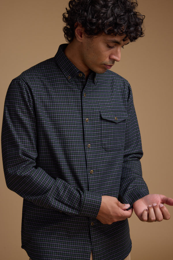 A man with curly hair looks down while buttoning the cuff of his Barkers Starle Check Shirt, made from responsibly sourced cotton, against a beige background.