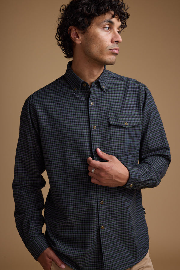 A man with curly hair wears the Barkers Starle Check Shirt, made from responsibly sourced cotton, and stands against a plain brown background. He looks to his left, holding the shirt near his chest with a neutral expression.
