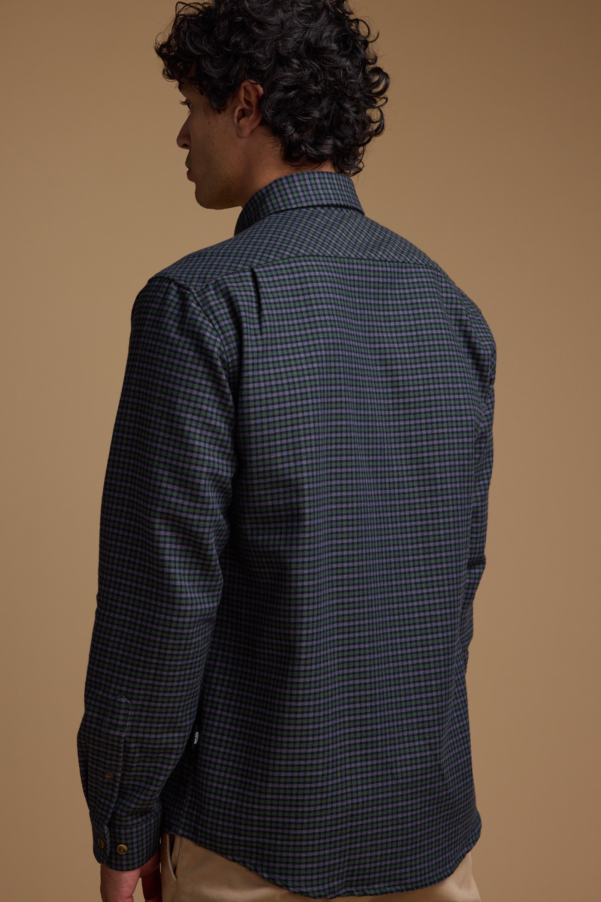 A man with curly dark hair stands with his back to the camera, wearing the Barkers Starle Check Shirt made from responsibly sourced cotton and beige pants, set against a plain brown background.