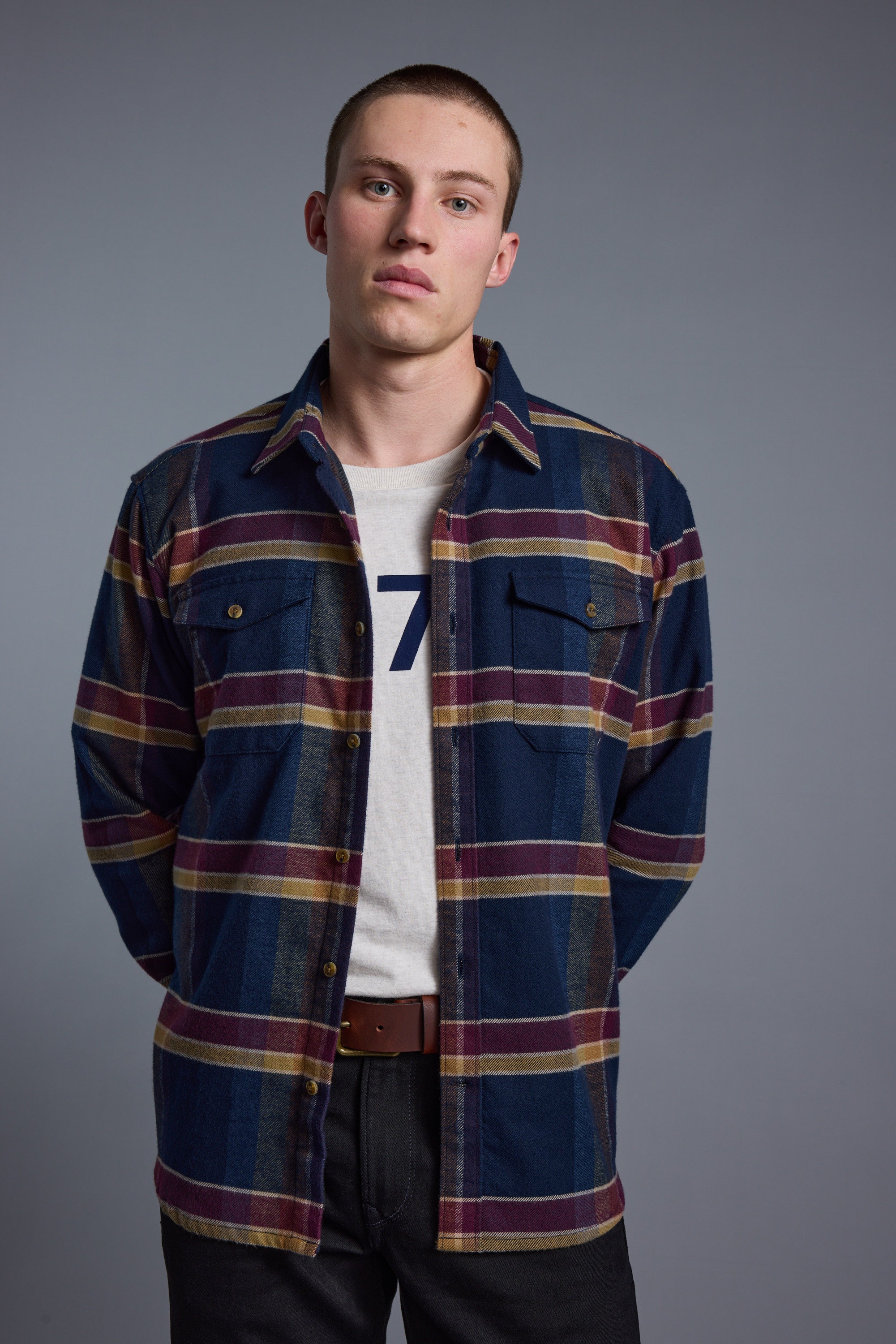 A young man stands against a plain background wearing the Barkers Heaton Check Overshirt—crafted from responsibly sourced cotton in vintage blue, red, and yellow plaid—layered over a white graphic tee. His hands are behind his back and he has a neutral expression.
