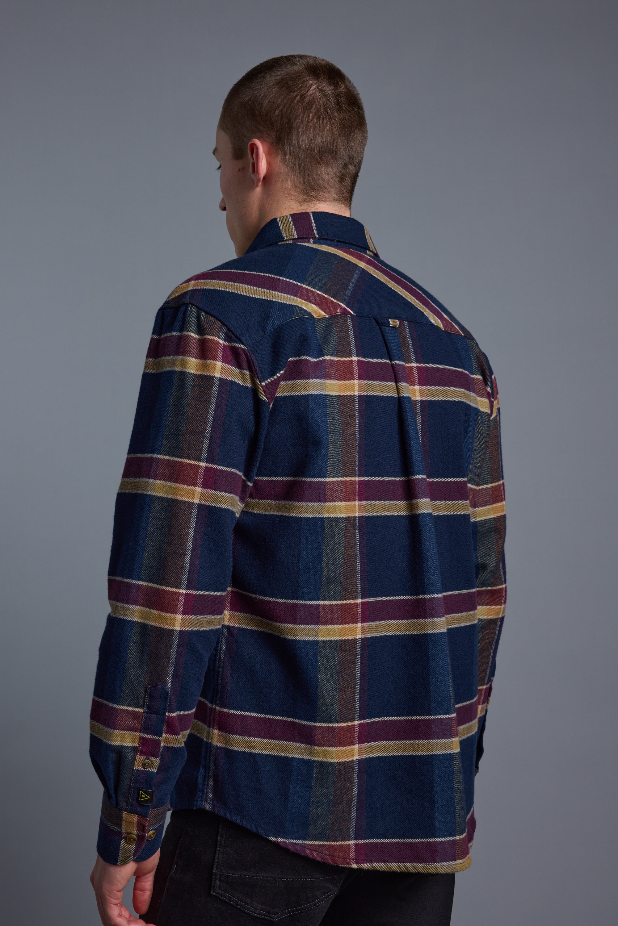 A person with short hair, seen from behind, wears the Barkers Heaton Check Overshirt in vintage dark blue, yellow, and maroon plaid with long sleeves, standing against a plain gray background.