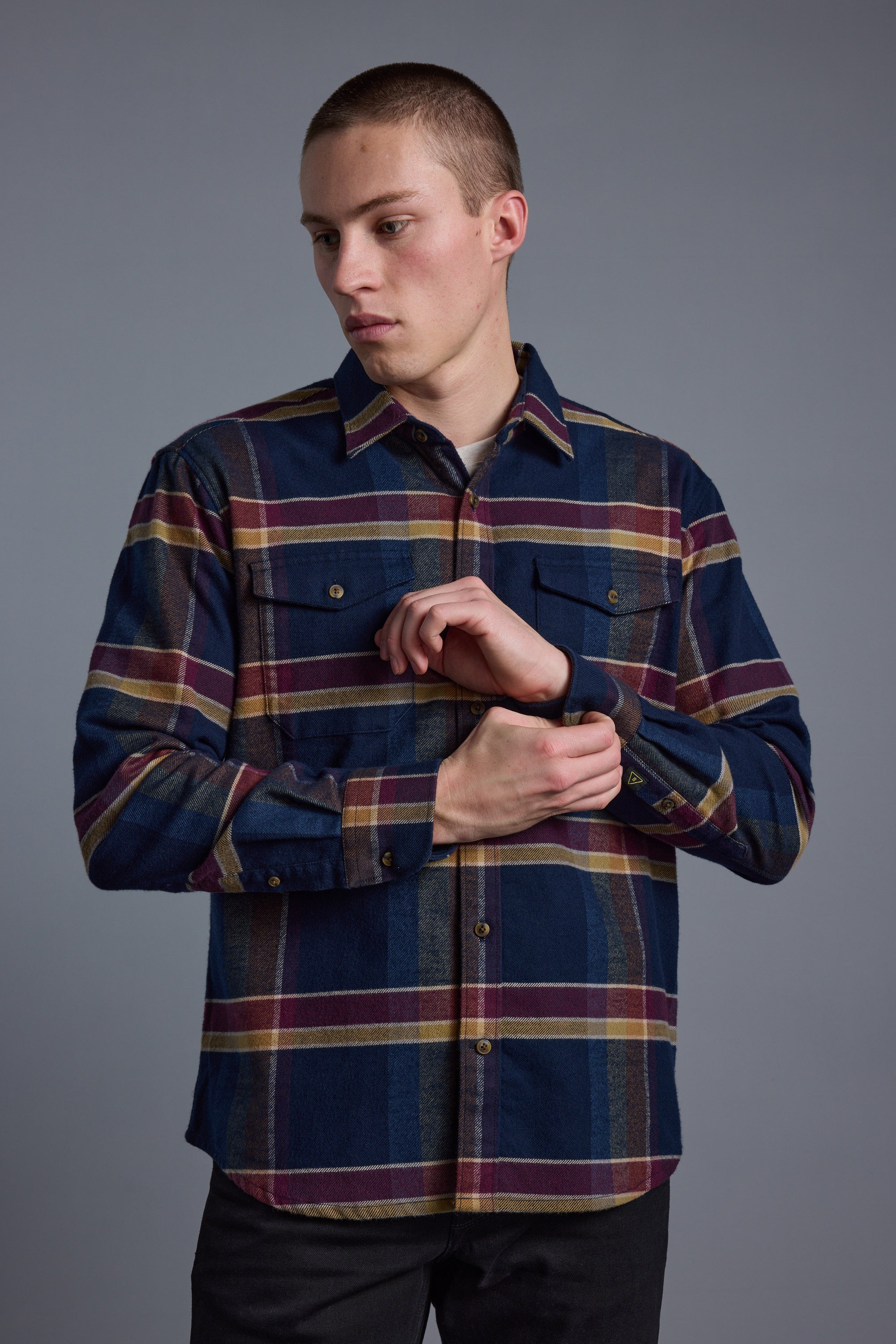 A young man in the Barkers Heaton Check Overshirt, crafted from responsibly sourced cotton, stands against a plain gray background and looks left while adjusting his sleeve.