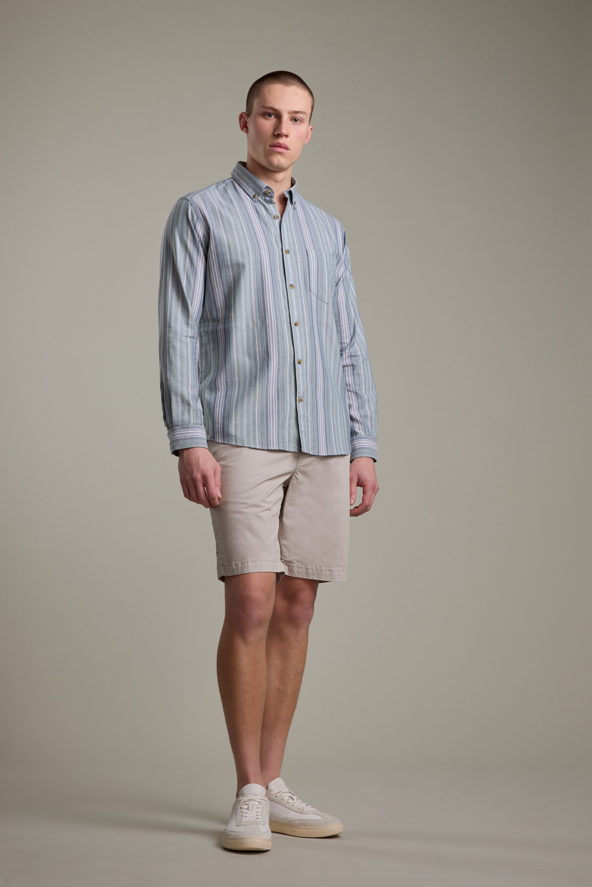 A man stands facing forward against a plain light gray background, wearing the Barkers Huntley Striped Oxford Shirt in blue and white stripes, paired with beige shorts and white sneakers.