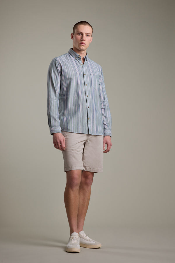 A man stands facing forward against a plain light gray background, wearing the Barkers Huntley Striped Oxford Shirt in blue and white stripes, paired with beige shorts and white sneakers.