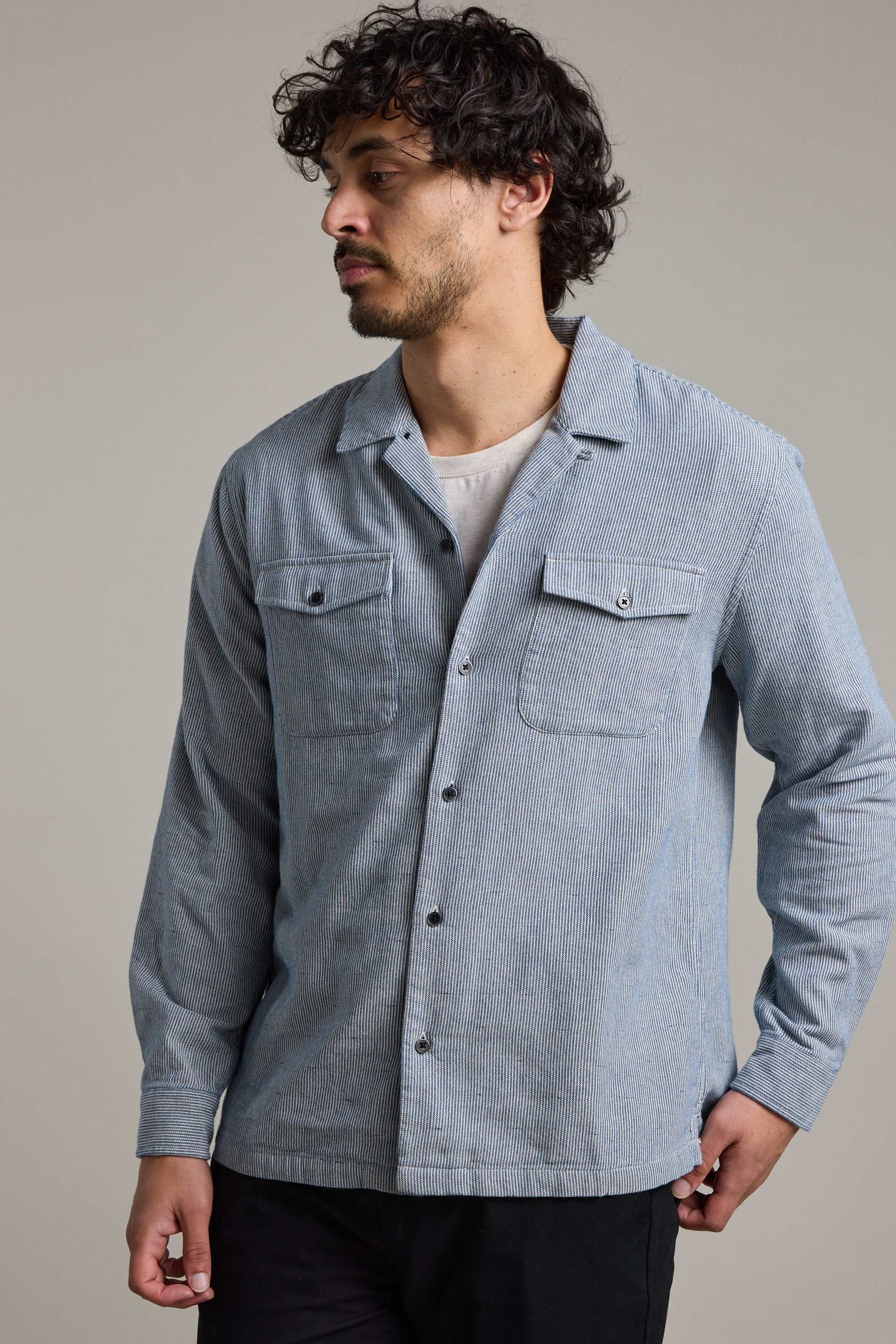 A man with curly hair and a beard wears the Barkers Sherman Cuban Overshirt in light blue over a white T-shirt, standing against a plain background and looking left with his right hand in his pocket.