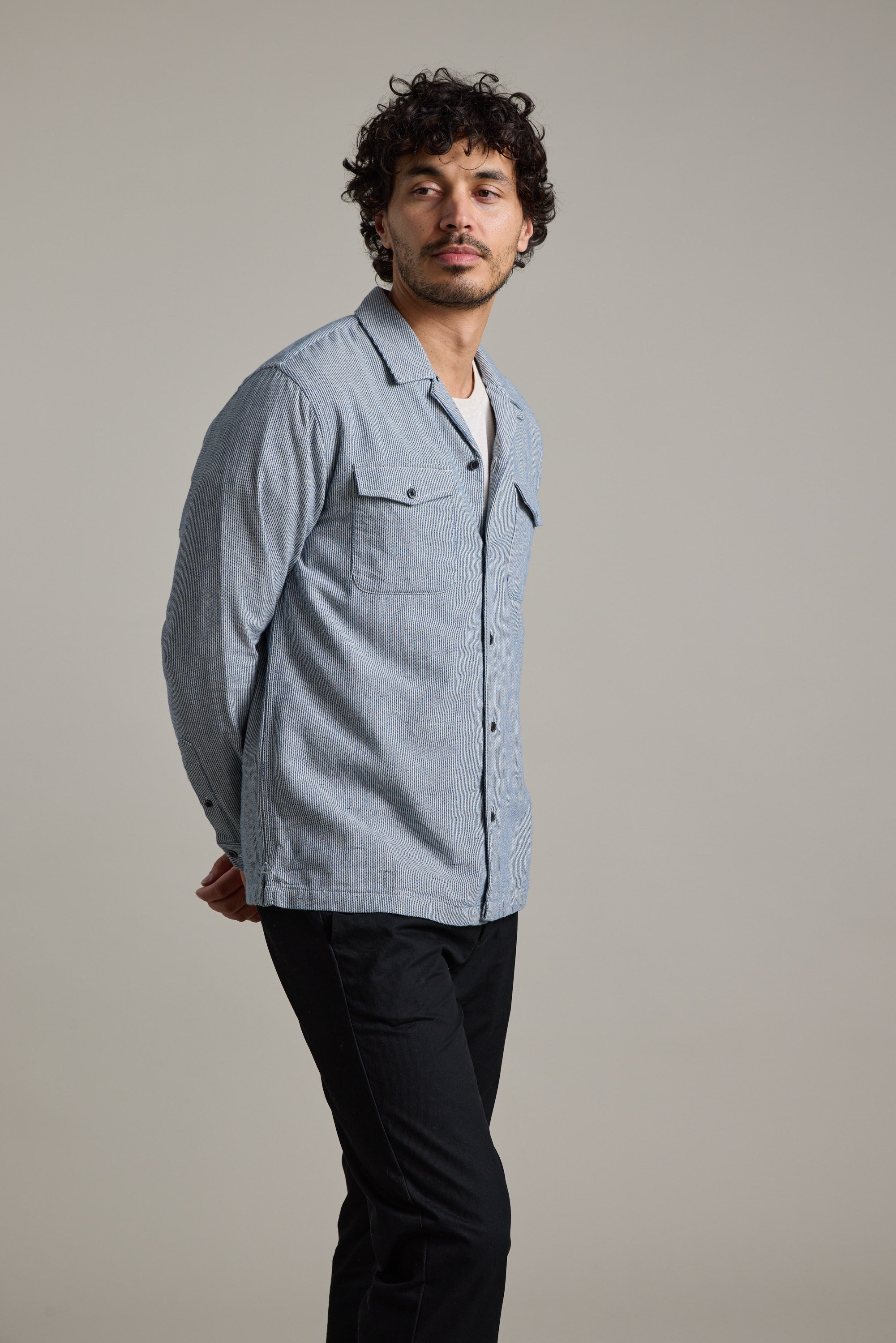 A man with curly hair models the Barkers Sherman Cuban Overshirt in light blue over a white t-shirt and black pants, standing hands behind his back and looking to the side against a plain background.