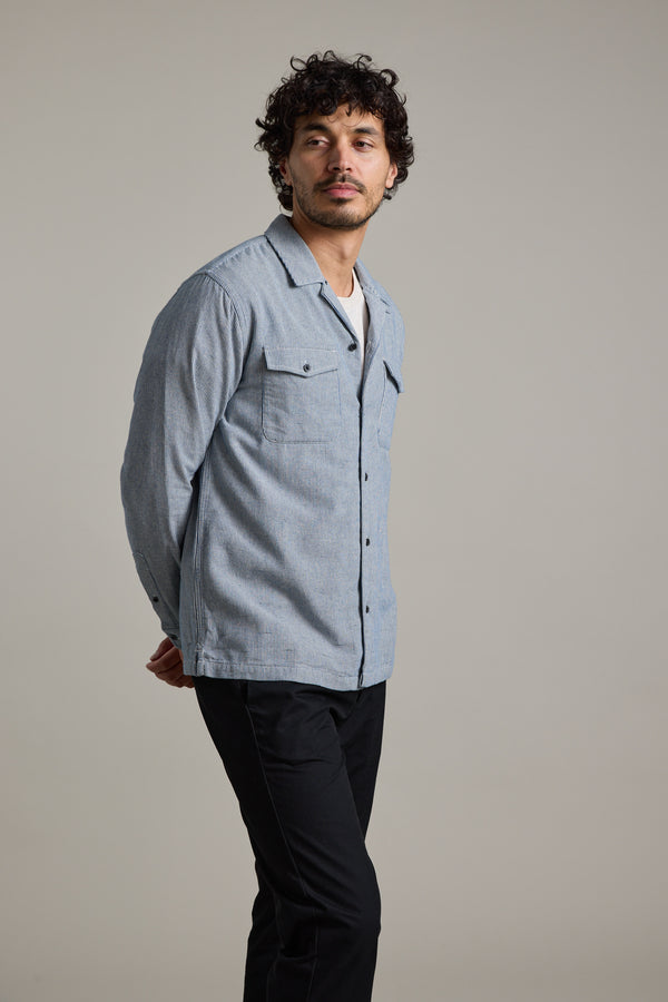 A man with curly hair models the Barkers Sherman Cuban Overshirt in light blue over a white t-shirt and black pants, standing hands behind his back and looking to the side against a plain background.