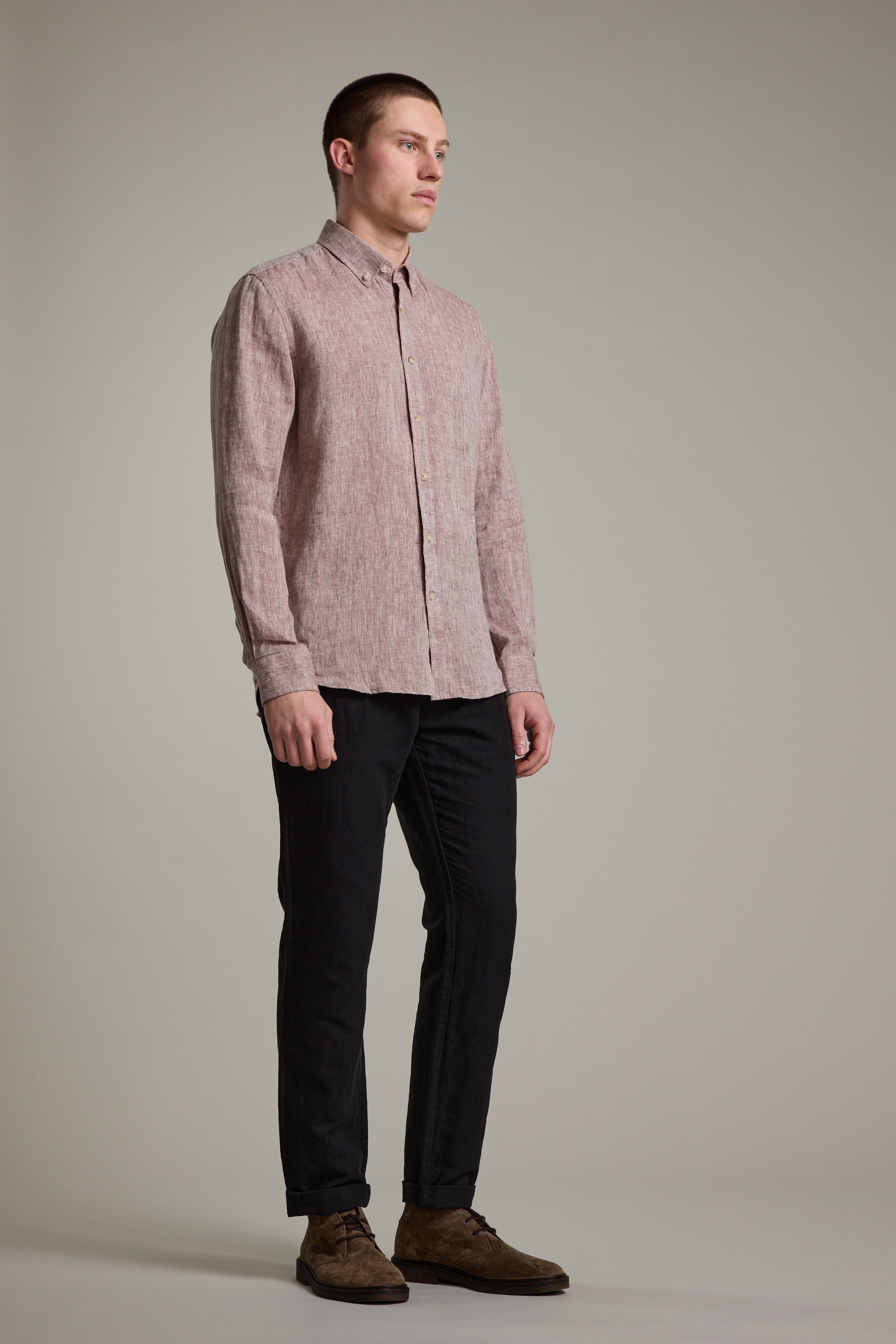 A man stands against a plain background, wearing the Barkers Dorset Linen Shirt in light pink with a tailored fit, paired with black pants and brown shoes. He faces left with a neutral expression, arms relaxed at his sides.