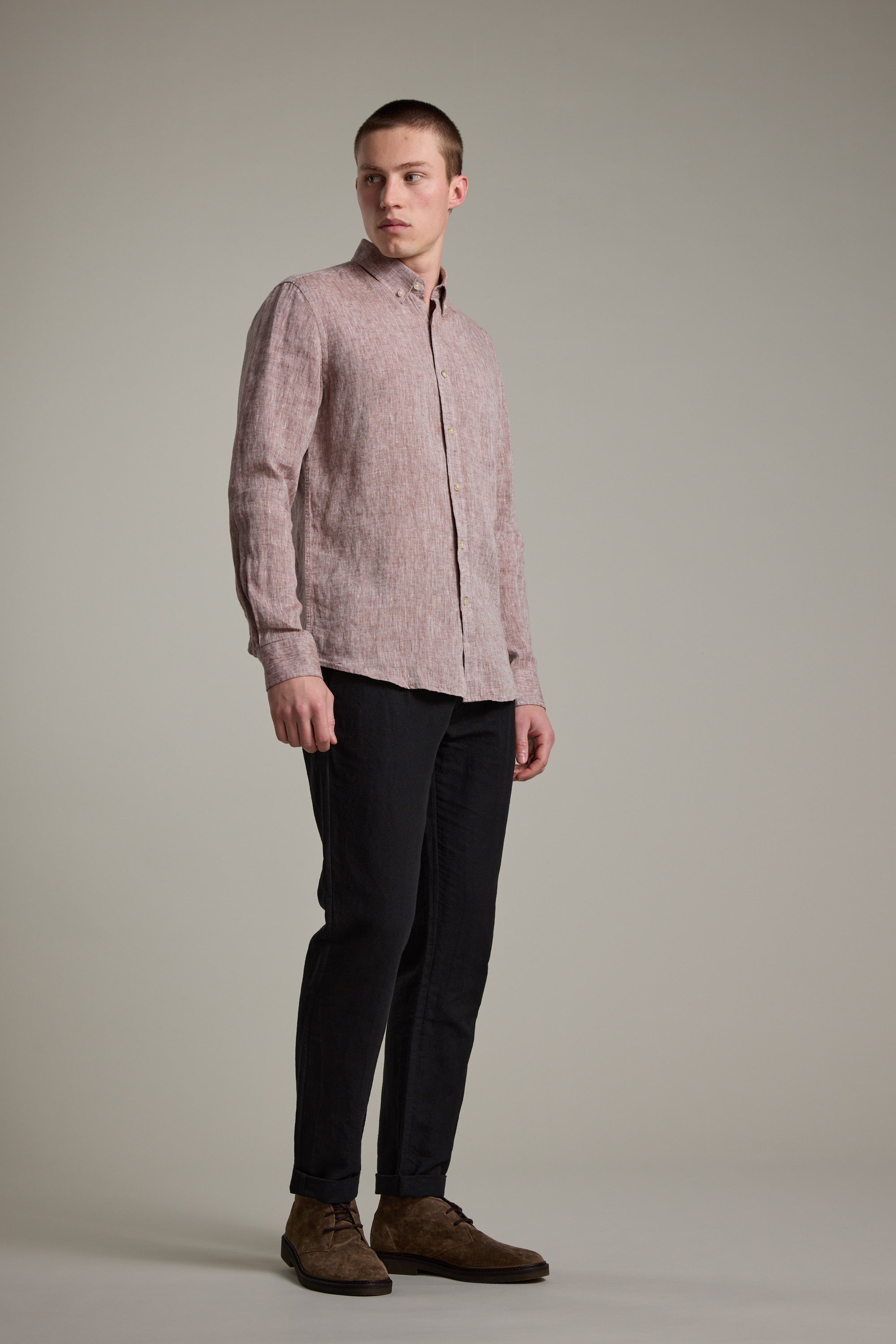 A man stands against a plain background wearing the Barkers Dorset Linen Shirt in light pink, featuring a tailored fit, paired with dark pants and brown shoes, gazing slightly to his left.