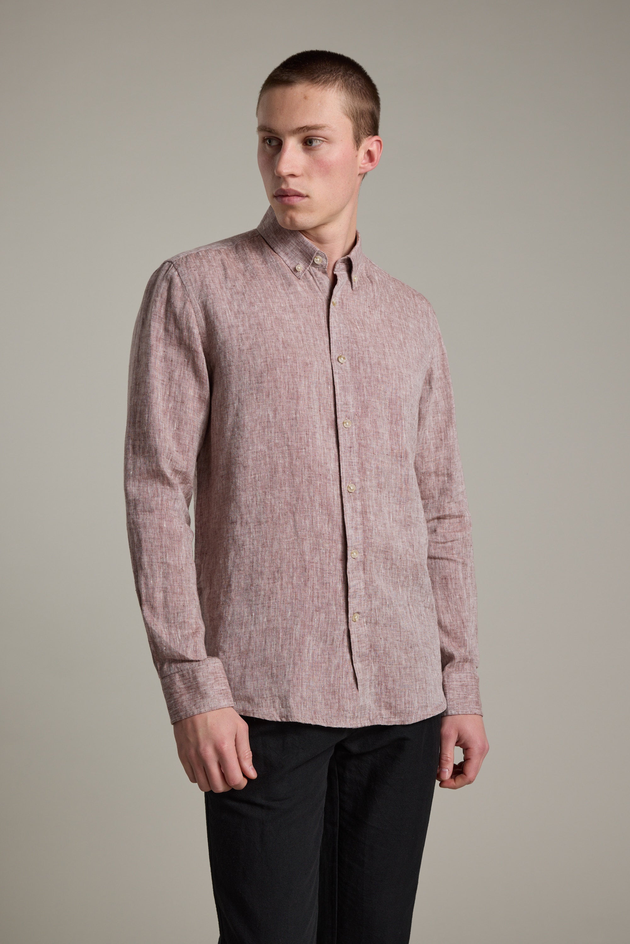 A young man wears the Barkers Dorset Linen Shirt in tailored fit, light red, paired with black pants, standing against a plain light gray background and looking slightly to his left.