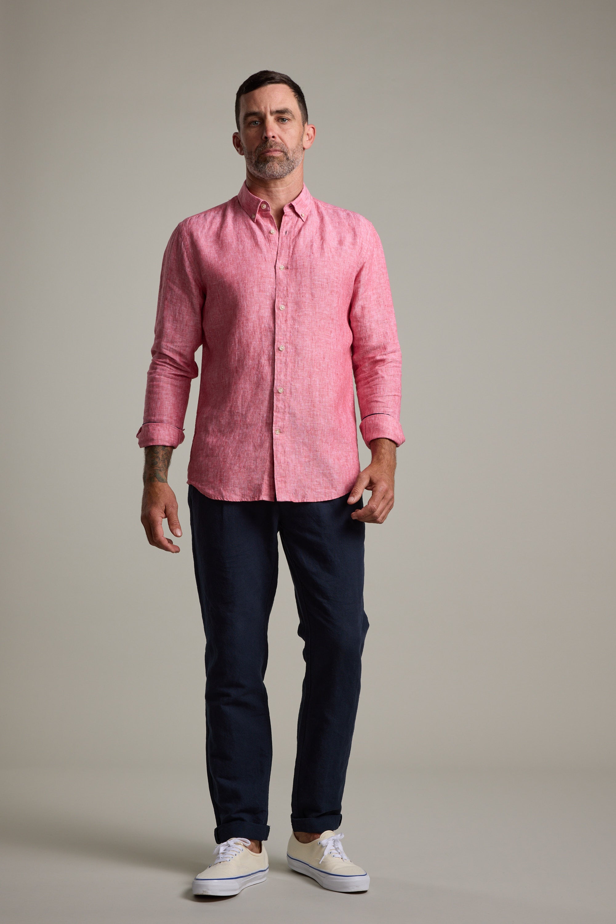 A man with short hair and a trimmed beard stands against a plain background wearing the Barkers Dorset Linen Shirt in pink, styled with dark pants and white sneakers. He looks straight ahead with a neutral expression.
