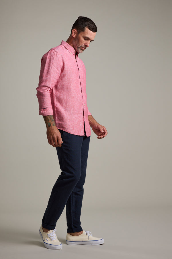 A man stands in profile against a plain background, wearing the Barkers Dorset Linen Shirt in pink, dark pants, and white sneakers. He looks downward with one leg slightly ahead.