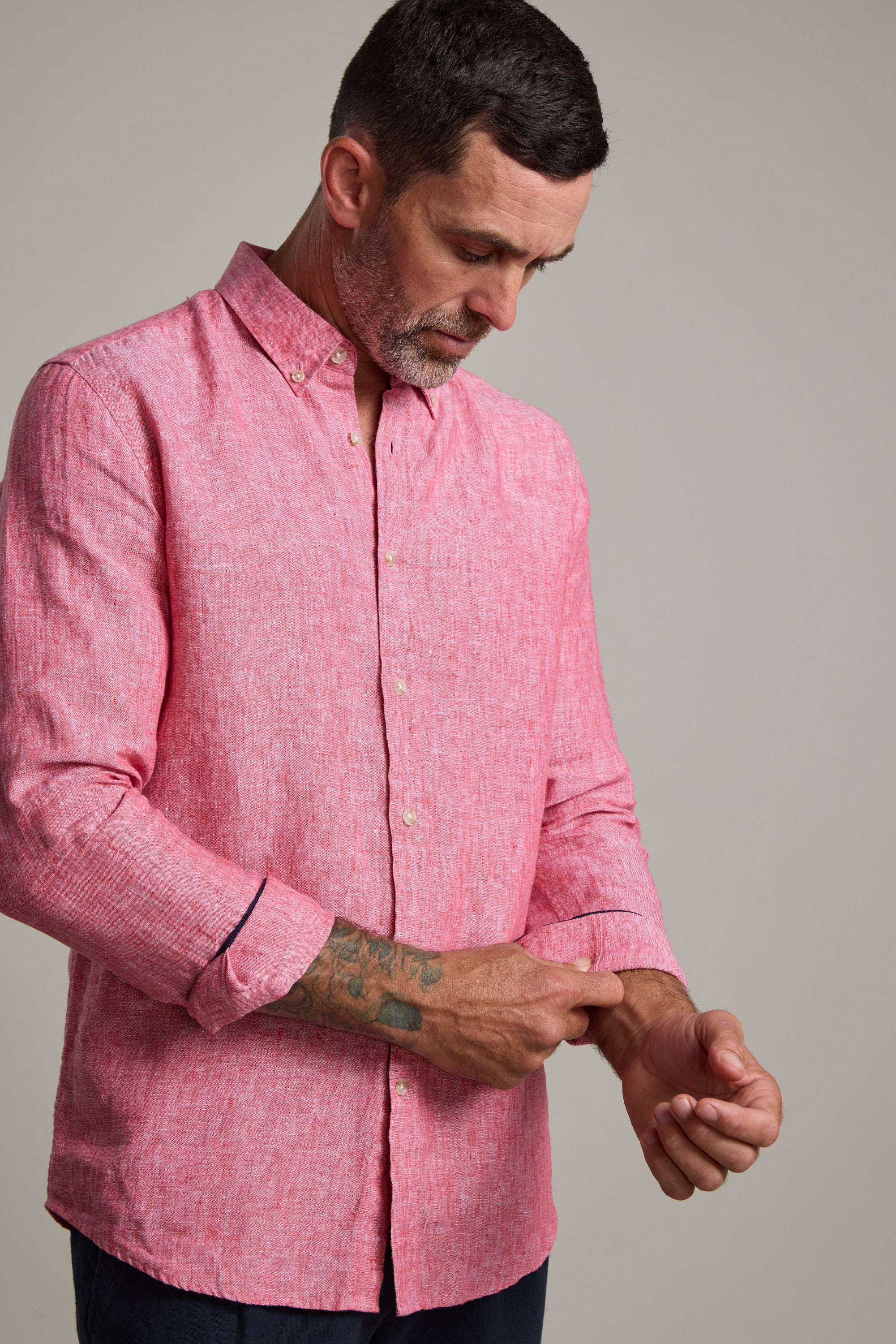 A man in a tailored fit Barkers Dorset Linen Shirt adjusts his cuff, revealing his tattooed left forearm. With short dark hair and a beard, he stands out in the breathable linen against a plain, neutral background.
