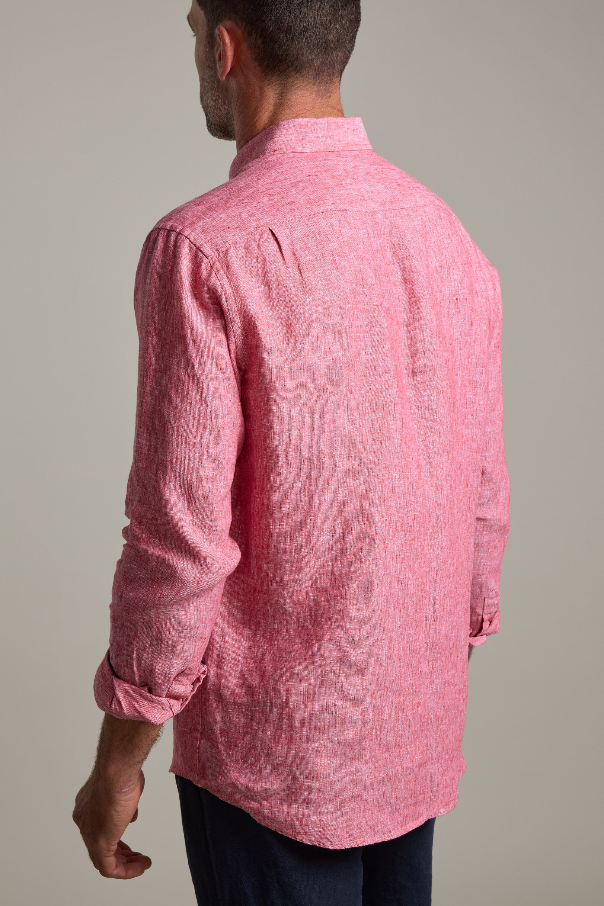 A man, seen from behind, wears the Barkers Dorset Linen Shirt in long-sleeve light red French linen with rolled-up sleeves and dark pants, standing against a plain, neutral background.