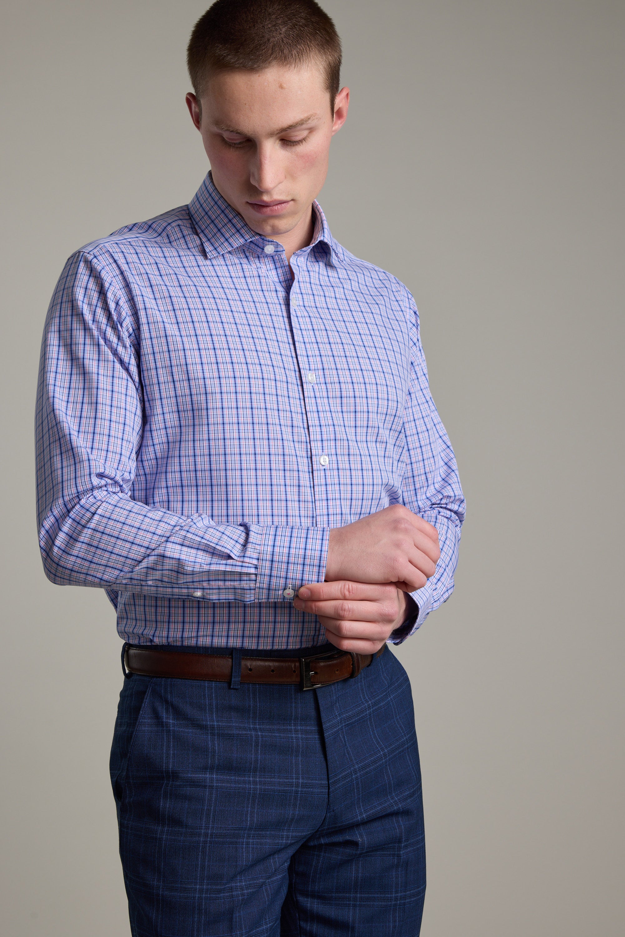 A man wearing the Barkers Denton Check Business Shirt in light purple adjusts his cuff while paired with blue plaid pants, against a plain neutral background.