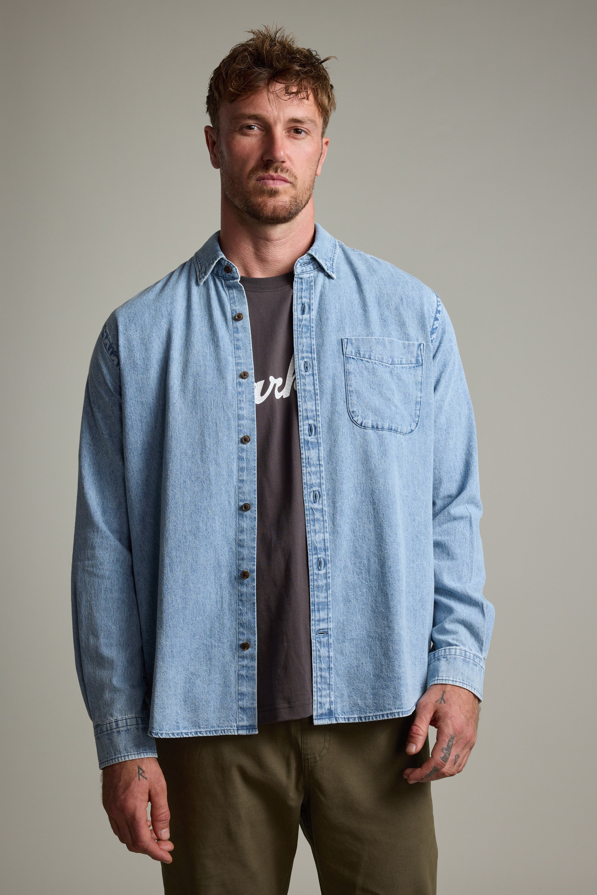 A man with short, tousled hair wears the Barkers Ruxton Denim Shirt as an overshirt over a black tee and olive green pants, standing against a plain gray background.