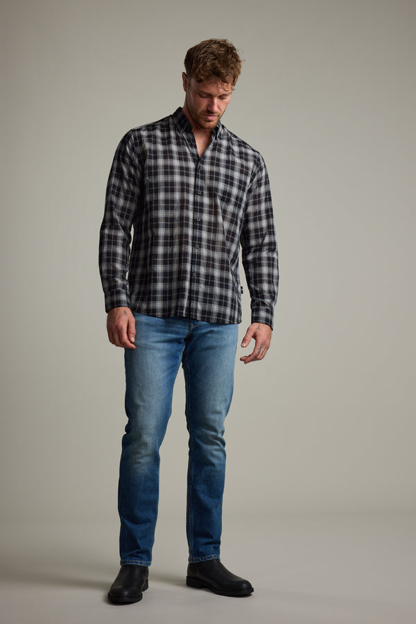 A man stands against a plain background, wearing the Barkers Carter Check Shirt in black and gray, paired with blue jeans and black shoes, looking down with his hands by his sides.
