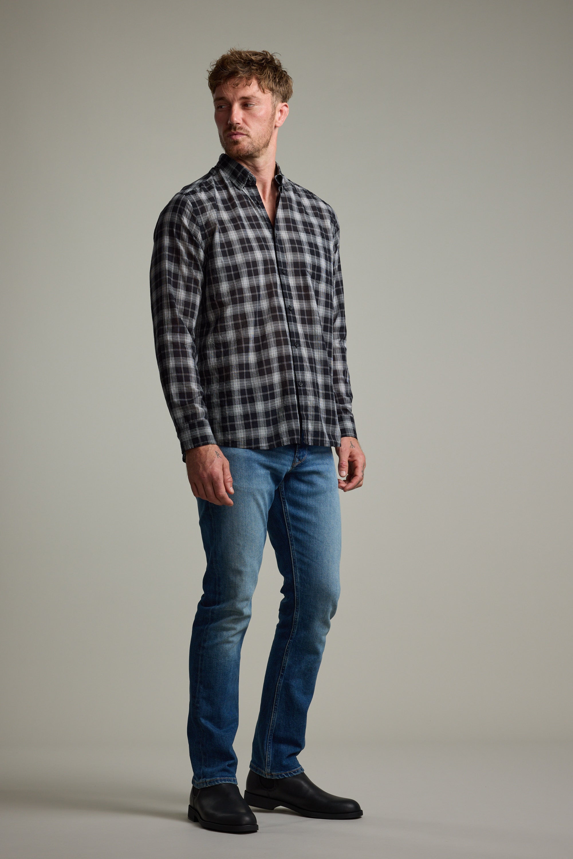 A man stands facing slightly left, wearing the Barkers Carter Check Shirt in black and white, paired with blue jeans and black boots, set against a plain light gray background.
