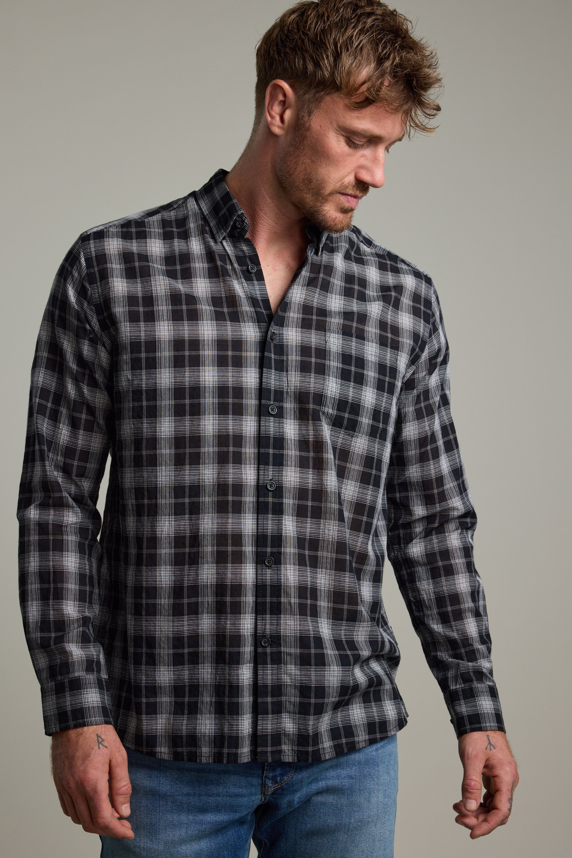 A man with short brown hair and light facial hair wears the Barkers Carter Check Shirt—a classic fit with a black, white, and grey check pattern—paired with blue jeans. He looks downward against a plain background.