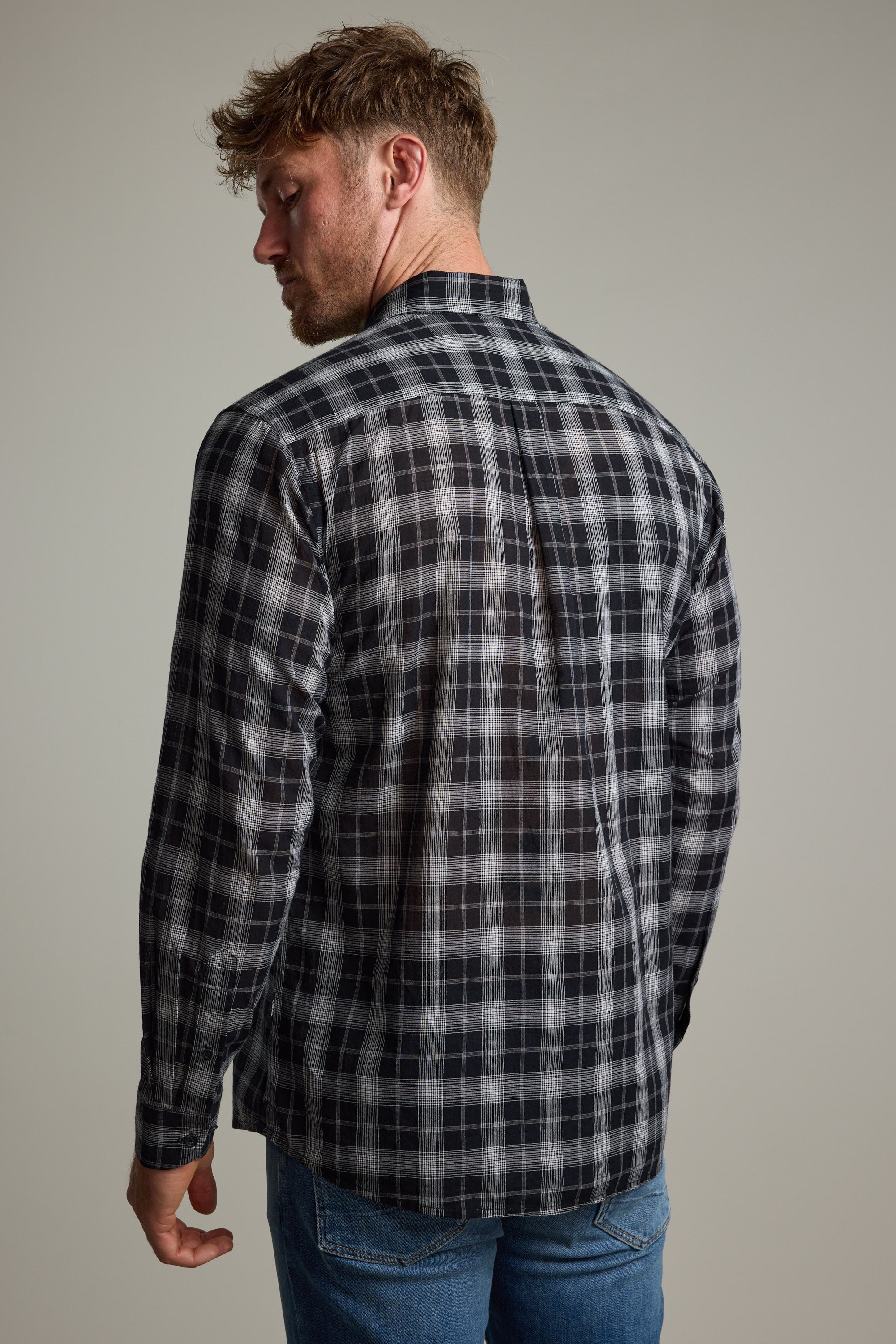A man with short, light brown hair stands with his back to the camera, wearing the Barkers Carter Check Shirt in black and gray, paired with blue jeans against a plain neutral background.