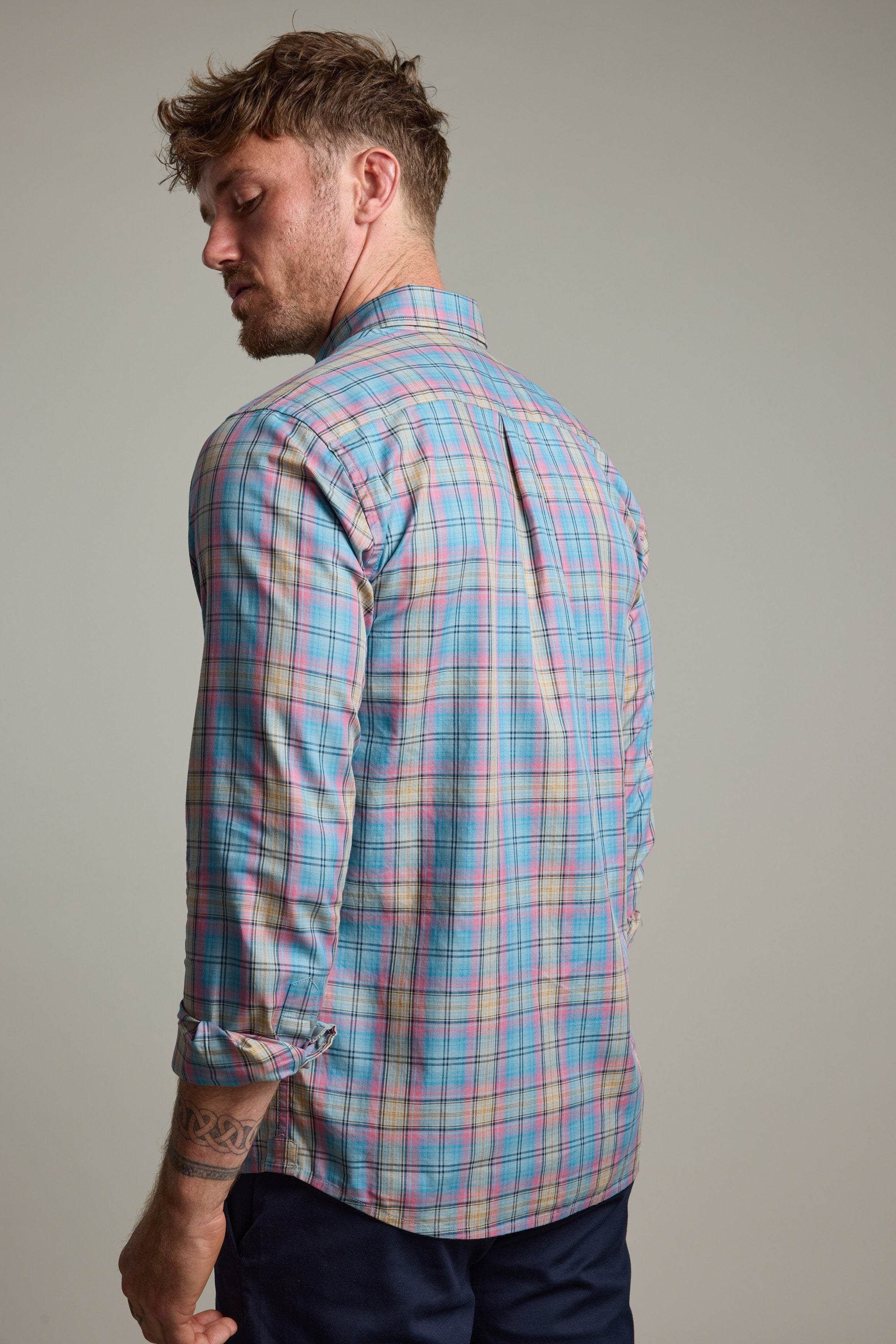A man with short, tousled hair stands facing away, wearing the Barkers Hayne Check Shirt in pastel plaid with rolled-up sleeves and dark pants. His tattooed left arm hangs loosely by his side.