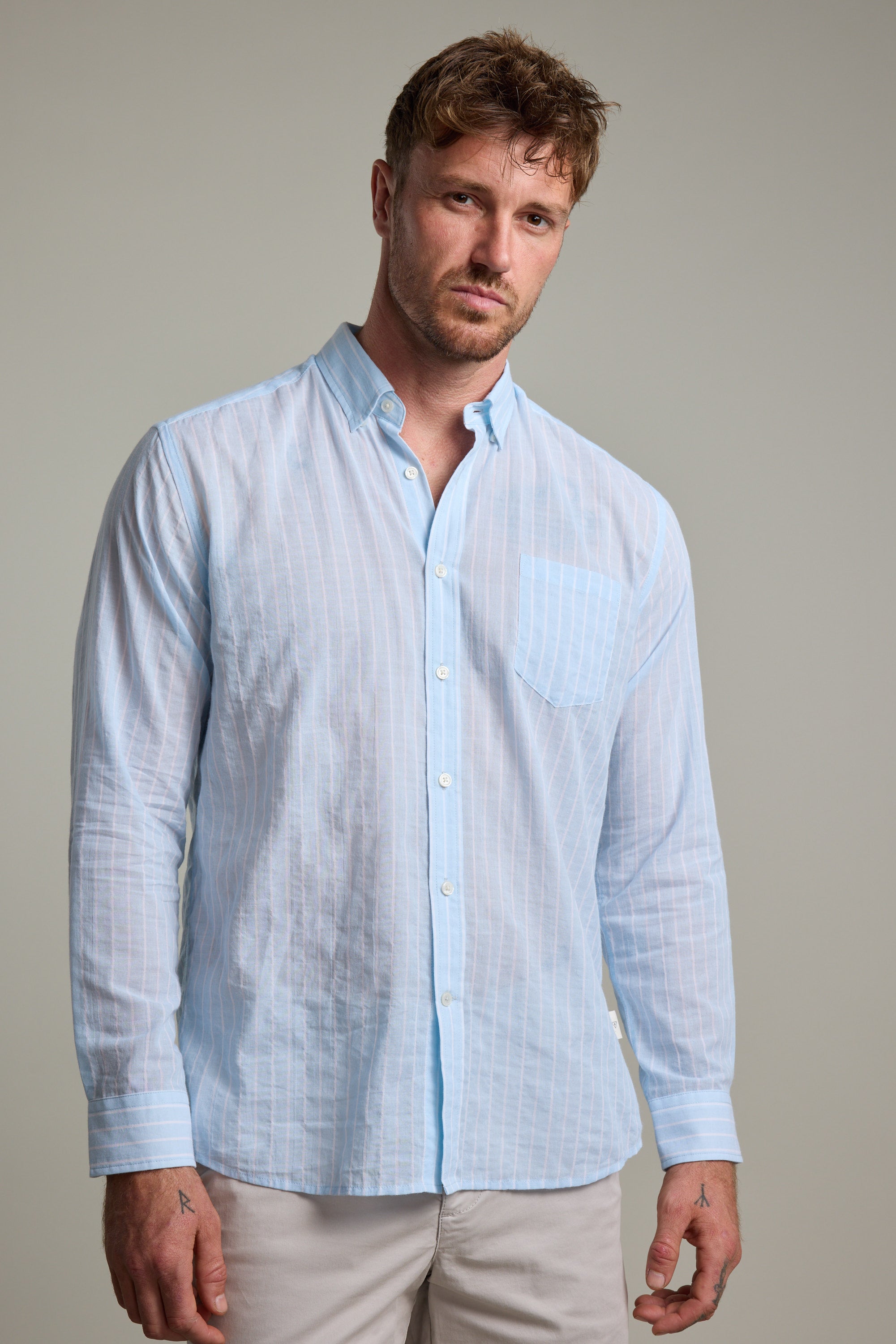 A man with short, tousled hair and a neutral expression wears the Barkers Russell Stripe Shirt—100% cotton, light blue and white stripes, with a pocket—standing against a plain light gray background.