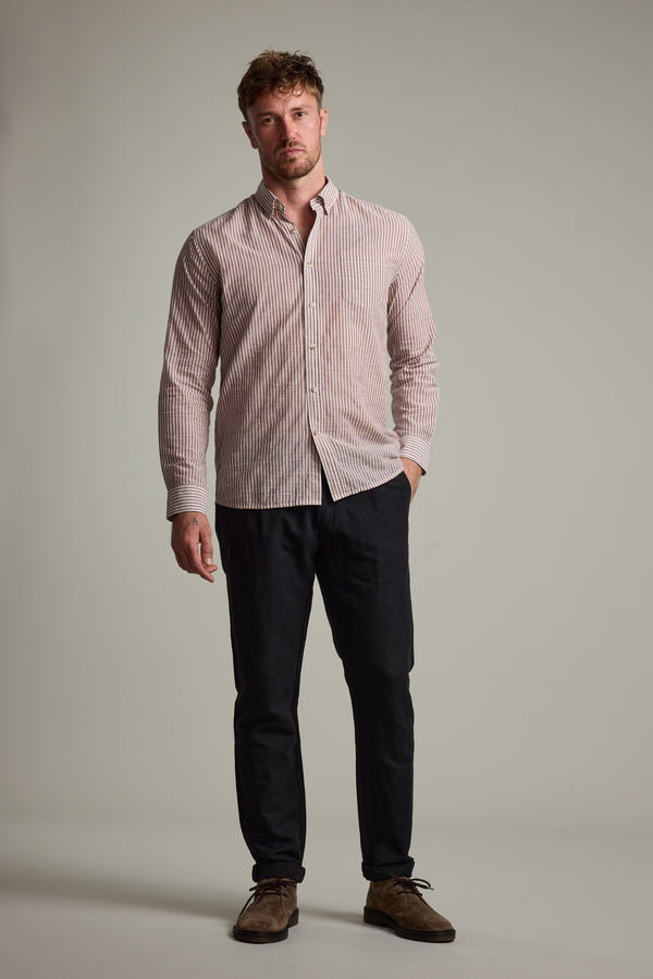 A man stands against a plain background wearing the Barkers Sumter Stripe Shirt in light pink and white, styled with dark pants and brown suede shoes, one hand casually tucked in his pocket.