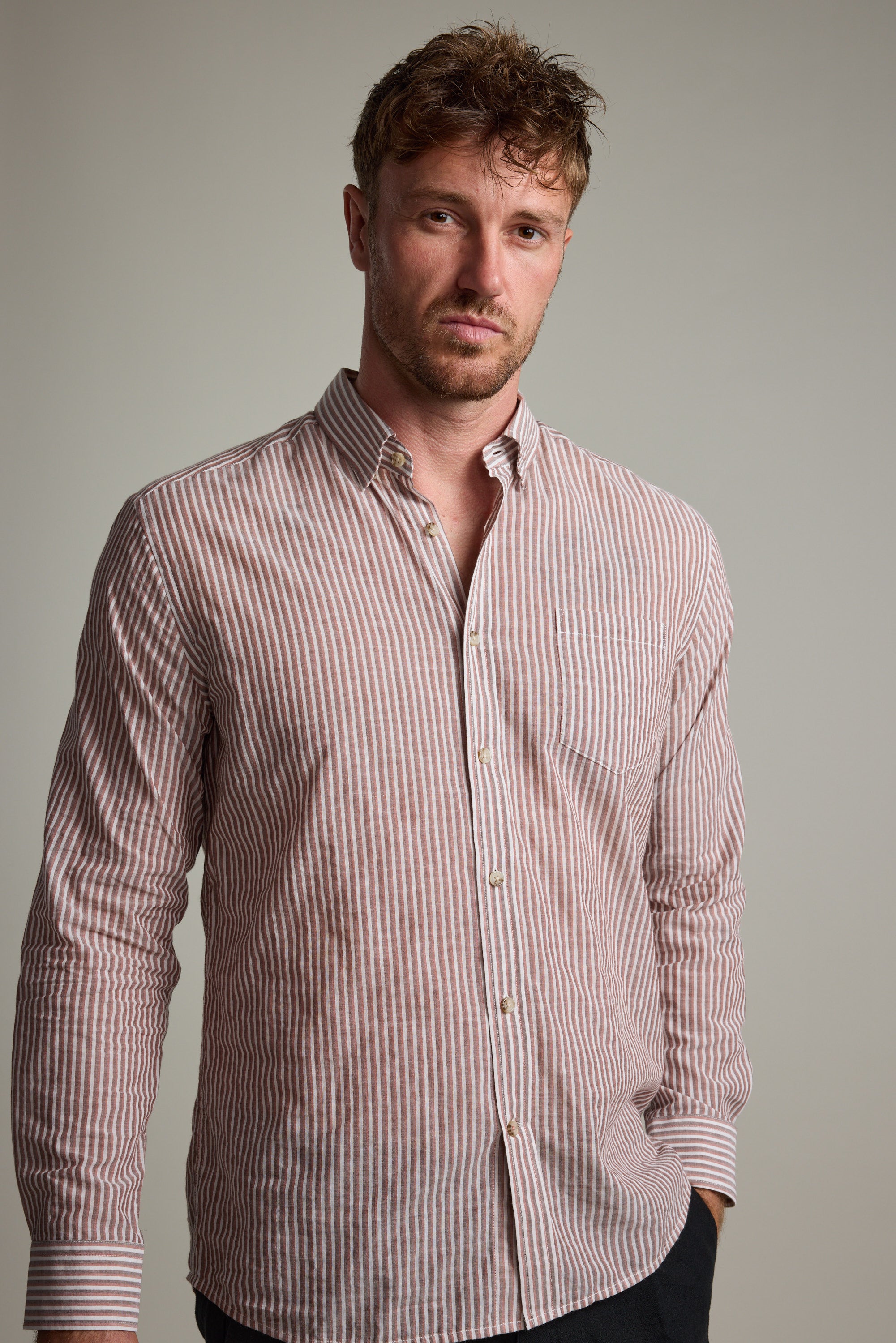 A man with short, tousled hair wears the Barkers Sumter Stripe Shirt, a lightweight long sleeve featuring vertical pink and white stripes. He stands against a plain light gray background, gazing neutrally at the camera.