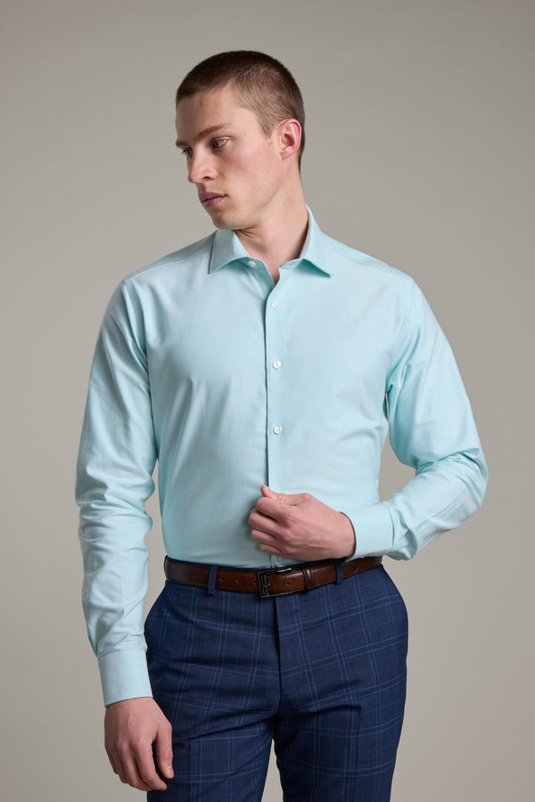 A man wearing the Barkers Edgar Texture Business Shirt in light blue and navy blue plaid pants stands against a plain background, looking to his left as he buttons his shirt with one hand.